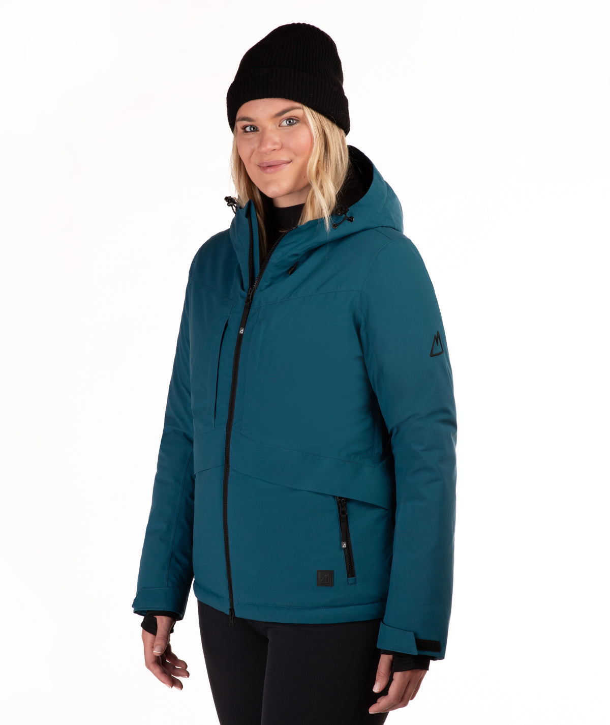 A woman wearing a teal ski jacket with black zippers, a black beanie, and black pants, photographed against a plain white studio background.