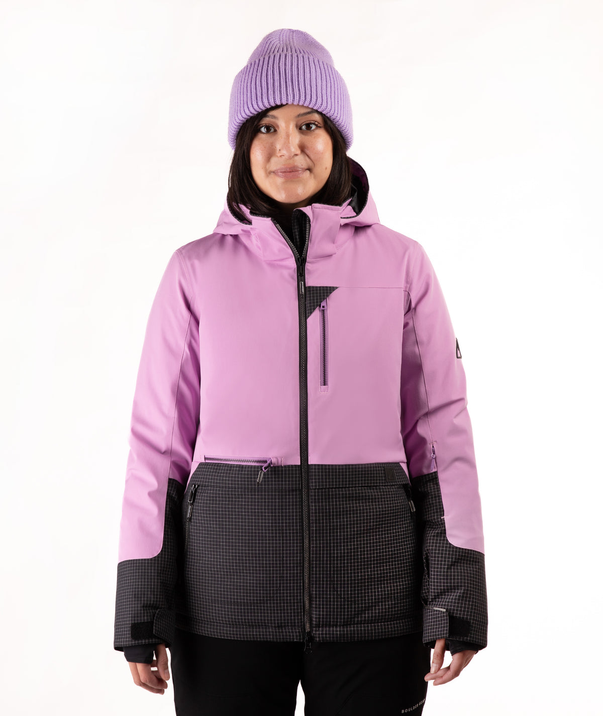 A woman wearing a light purple and black check ski jacket with a matching purple knit beanie, photographed against a plain white studio background.