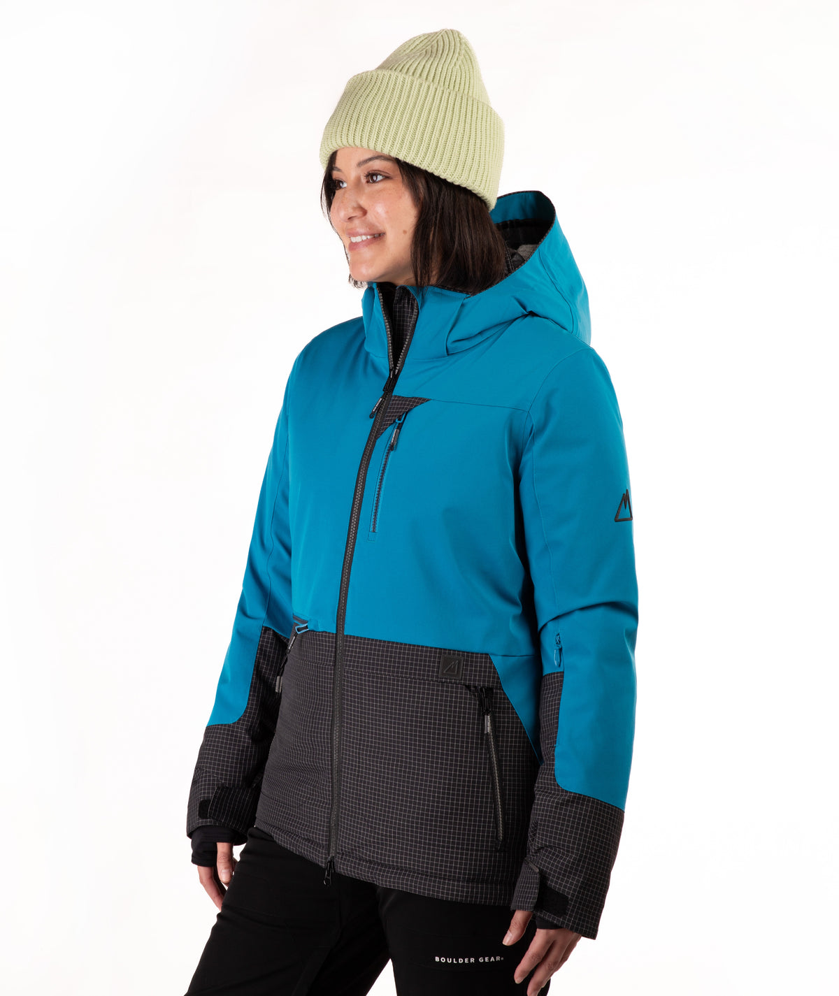 A woman wearing a bright turquoise and black check ski jacket, a light green ribbed beanie, and black snow pants, photographed against a clean white studio background.