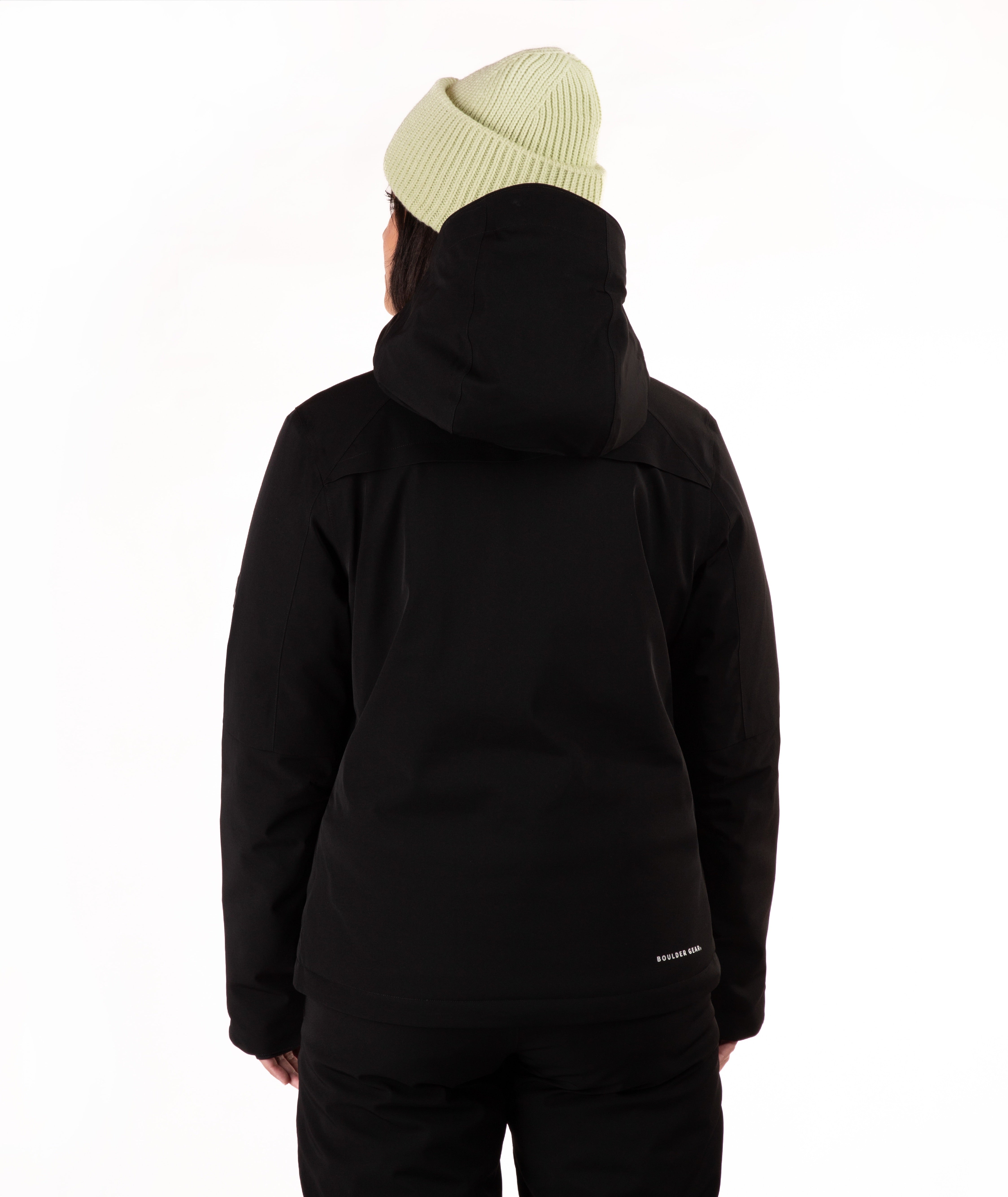 A woman wearing a black ski jacket and matching black pants, paired with a light green knit beanie, standing against a plain white studio background.