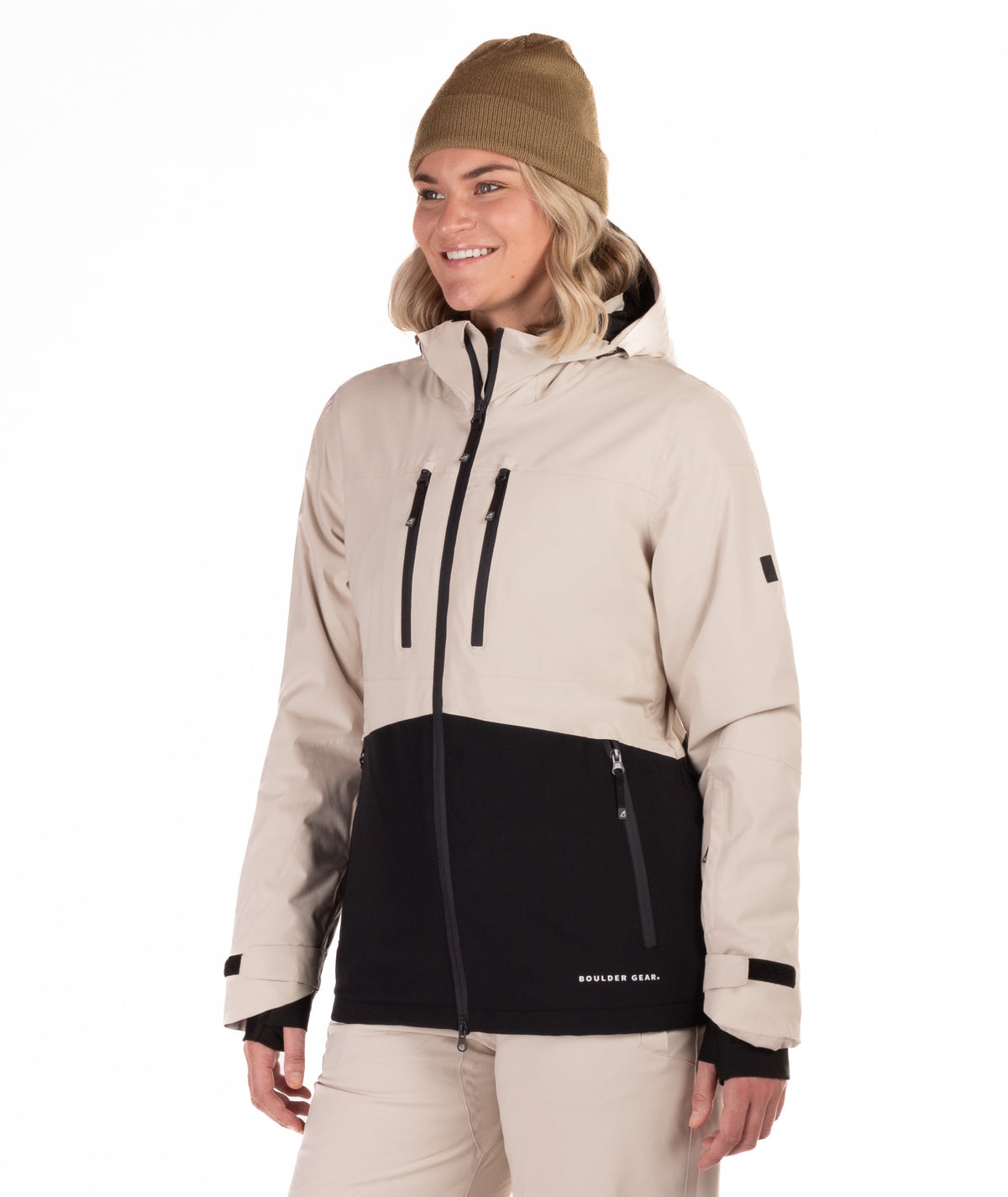 A woman wearing a beige and black snowboard jacket, a tan beanie, and beige snow pants, standing against a solid white background.