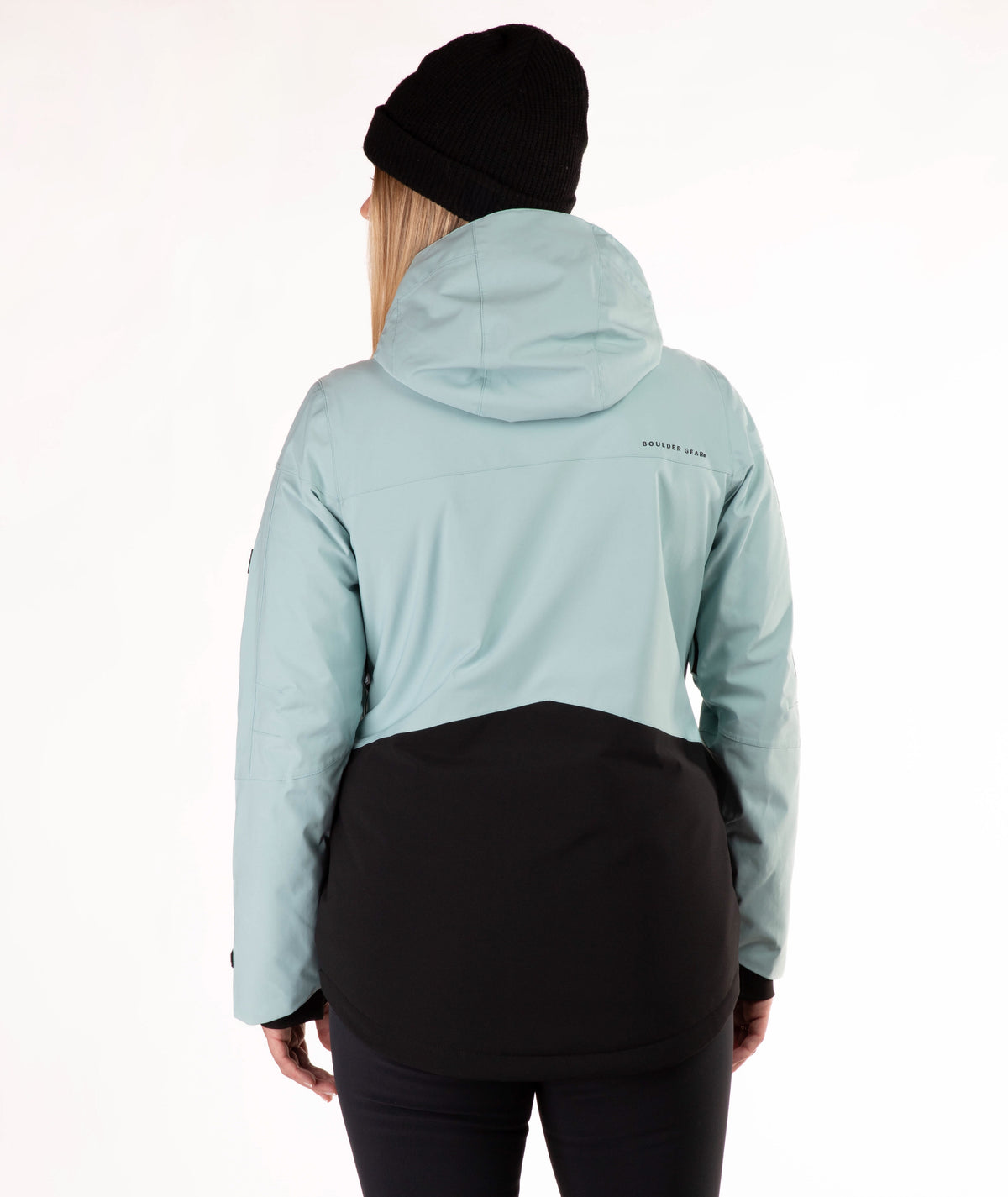 A woman wearing a light green and black ski jacket, a black beanie, and black pants, standing against a plain white studio background.