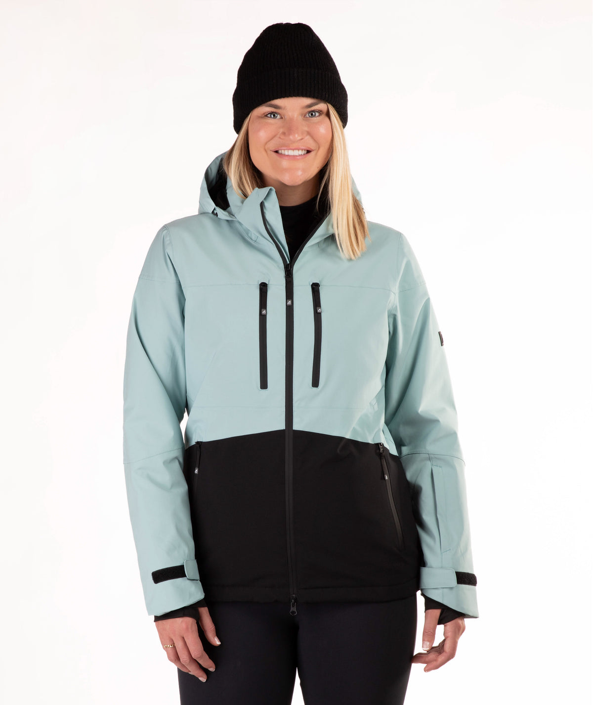 A woman wearing a light green and black ski jacket, a black beanie, and black pants, standing against a plain white studio background.