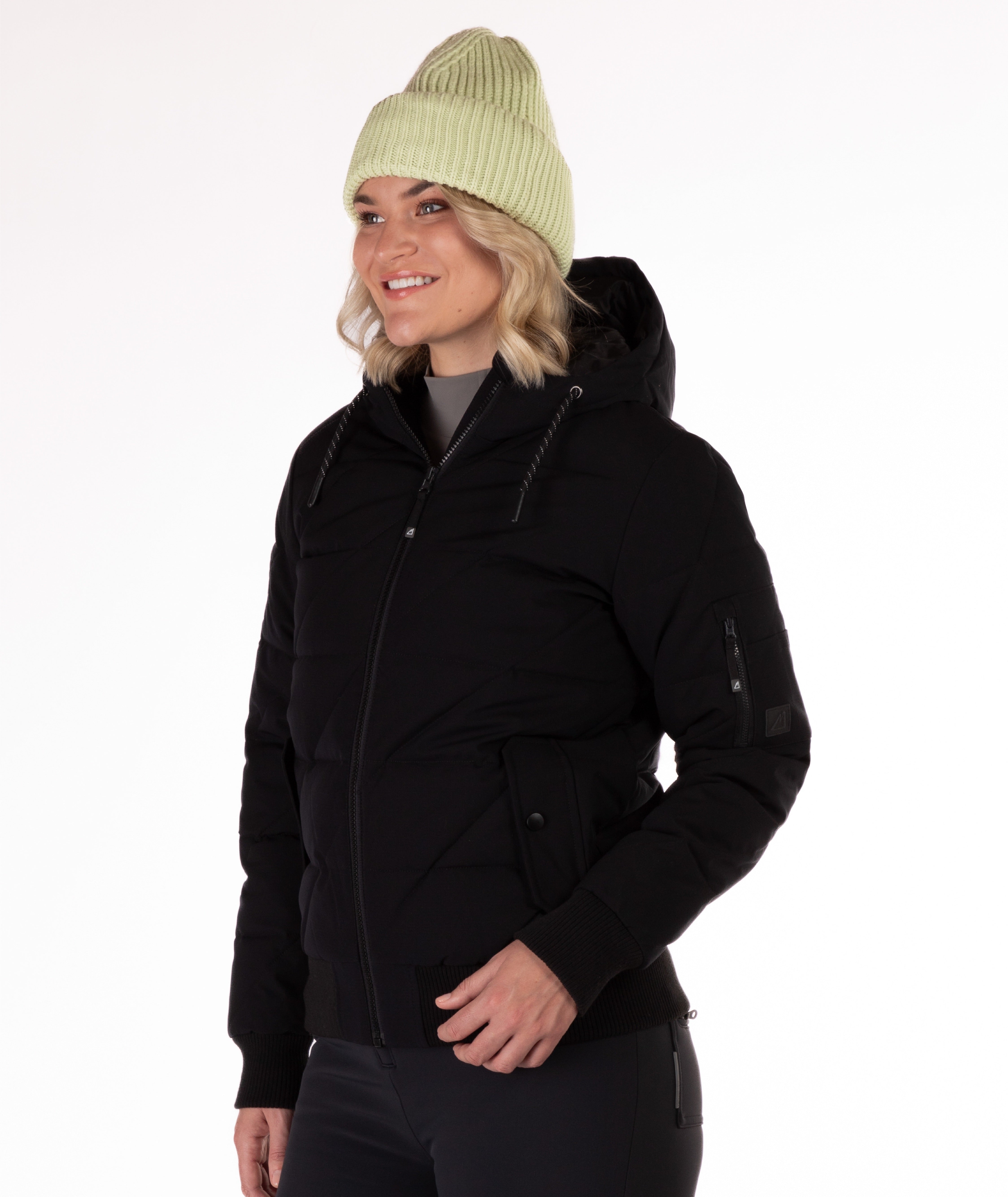 A woman wearing a black quilted jacket with a hood and black knit trim on wrist cuffs and hem on a white background. Side Profile view of the garment highlighting a cargo pocket on the left sleeve.