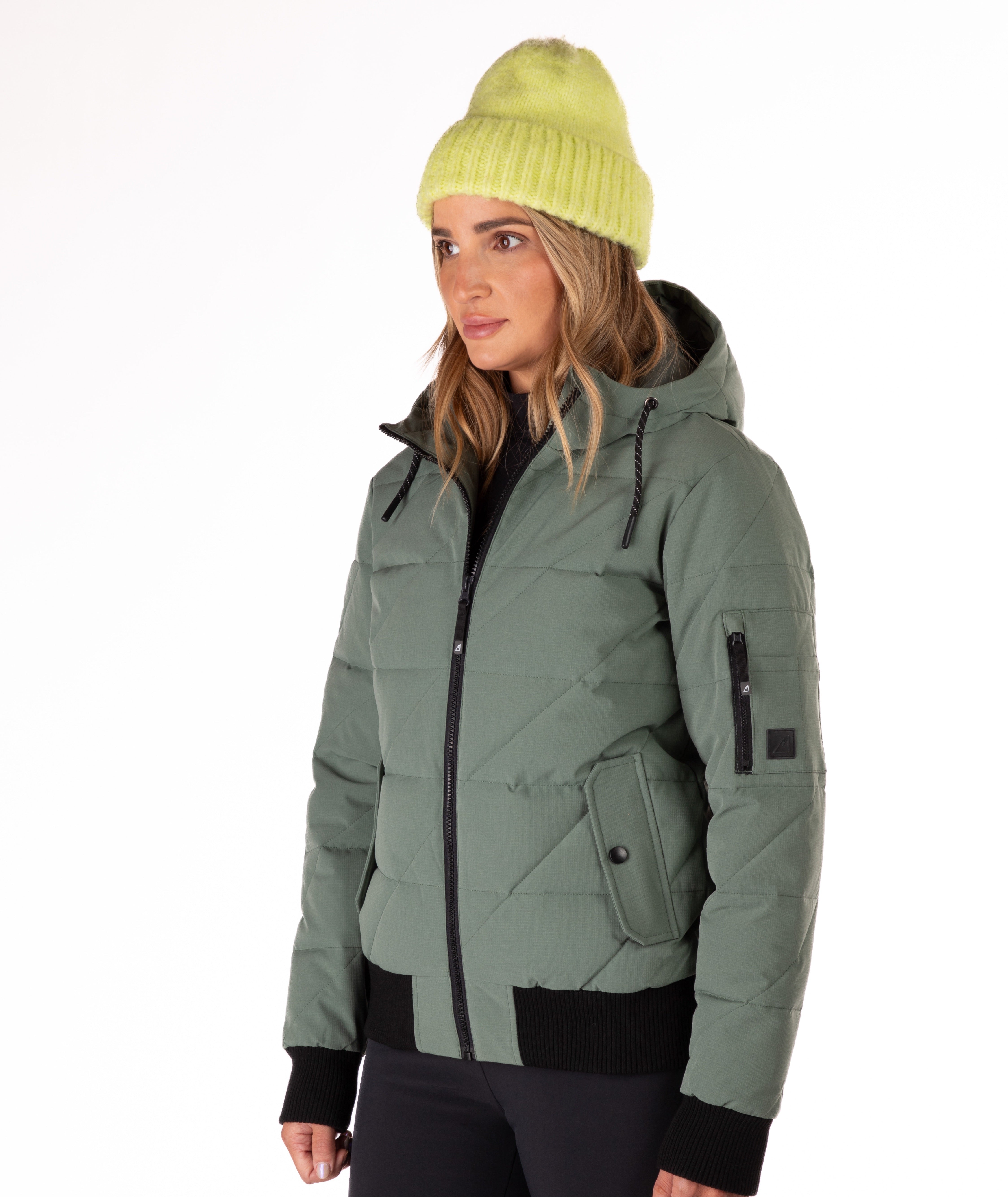Woman wearing a green quilted jacket with a hood and black knit trim on wrist cuffs and hem on a white background. Side profile view of the jacket highlighting a cargo pocket on the left sleeve.