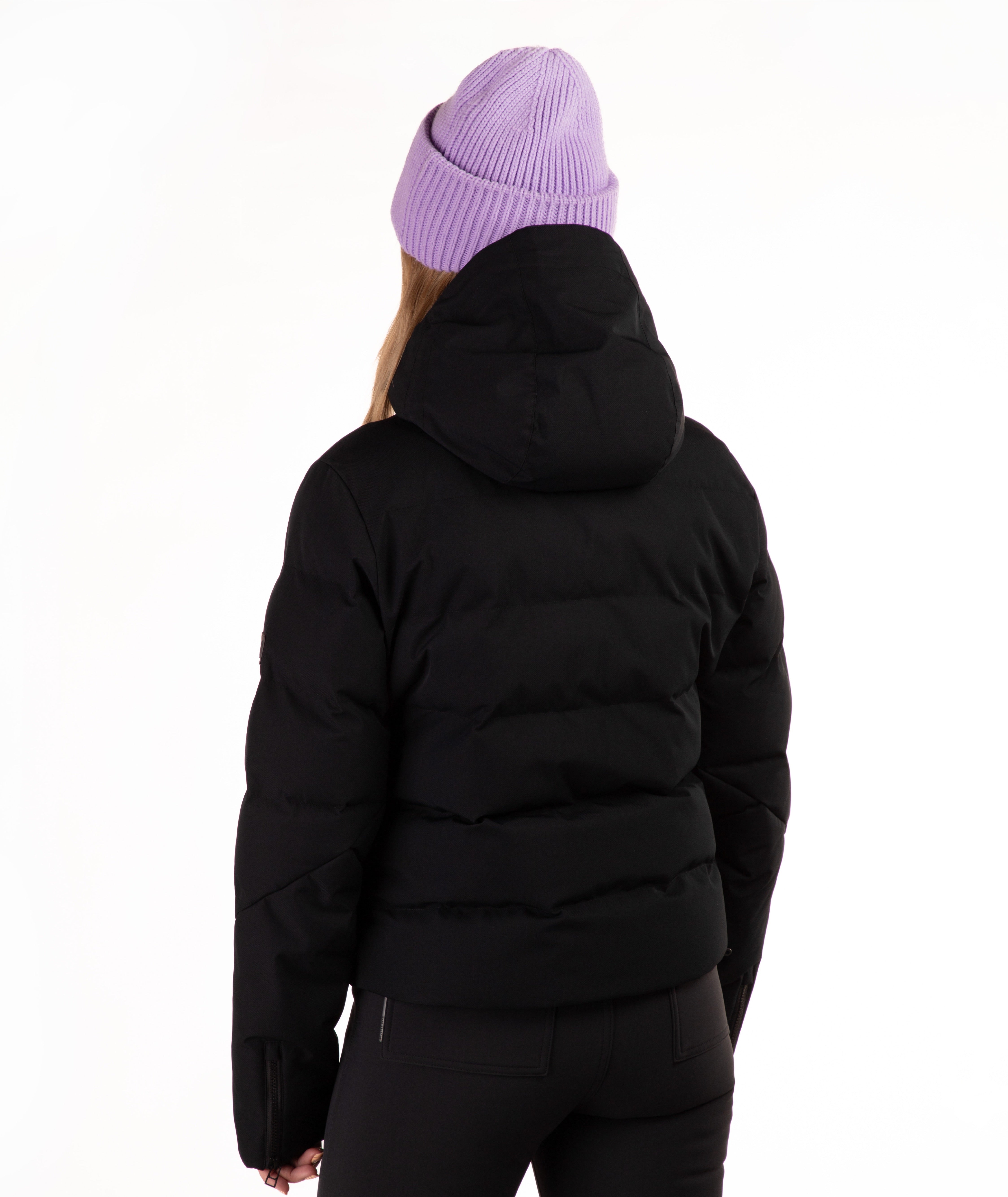 A person shown from the back wearing a black hooded puffer jacket, black pants, and a light purple ribbed knit beanie against a plain white studio background.