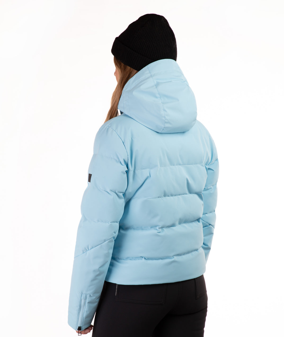 A person shown from the back wearing a light blue hooded puffer jacket, black beanie, and black pants against a plain white studio background.
