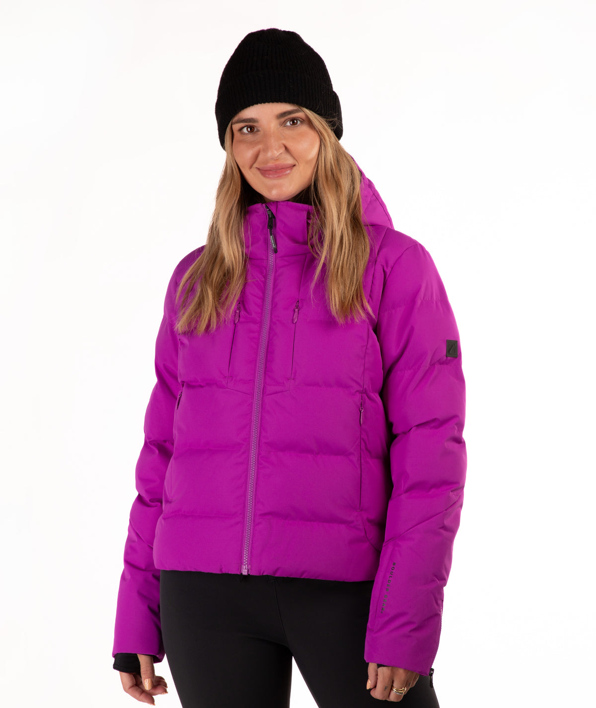 A woman wearing a bright magenta puffer jacket, a black beanie, and black pants, standing against a plain white studio background.