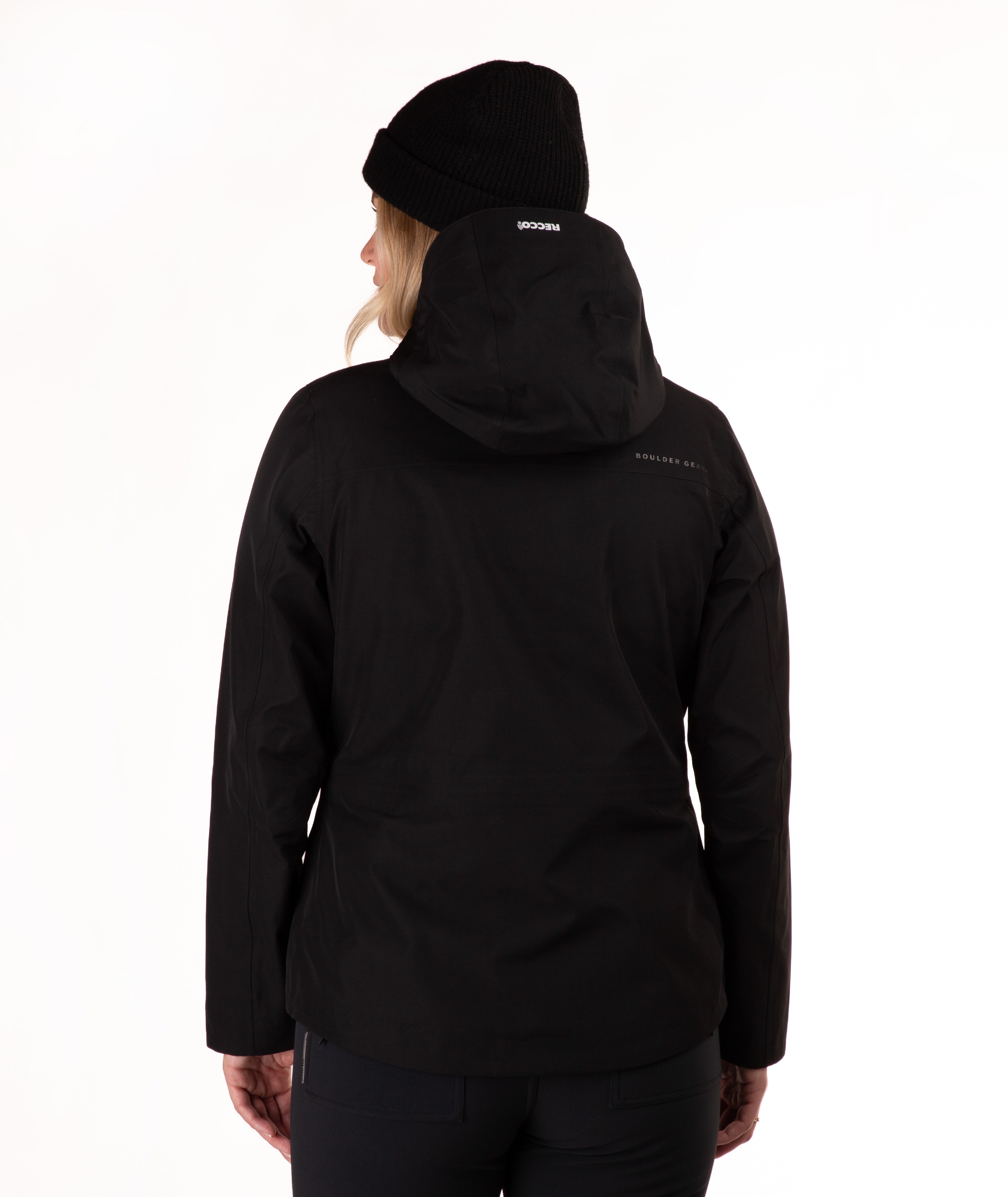 A woman viewed from the back wearing a black hooded jacket and a black beanie, photographed against a clean white studio background.
