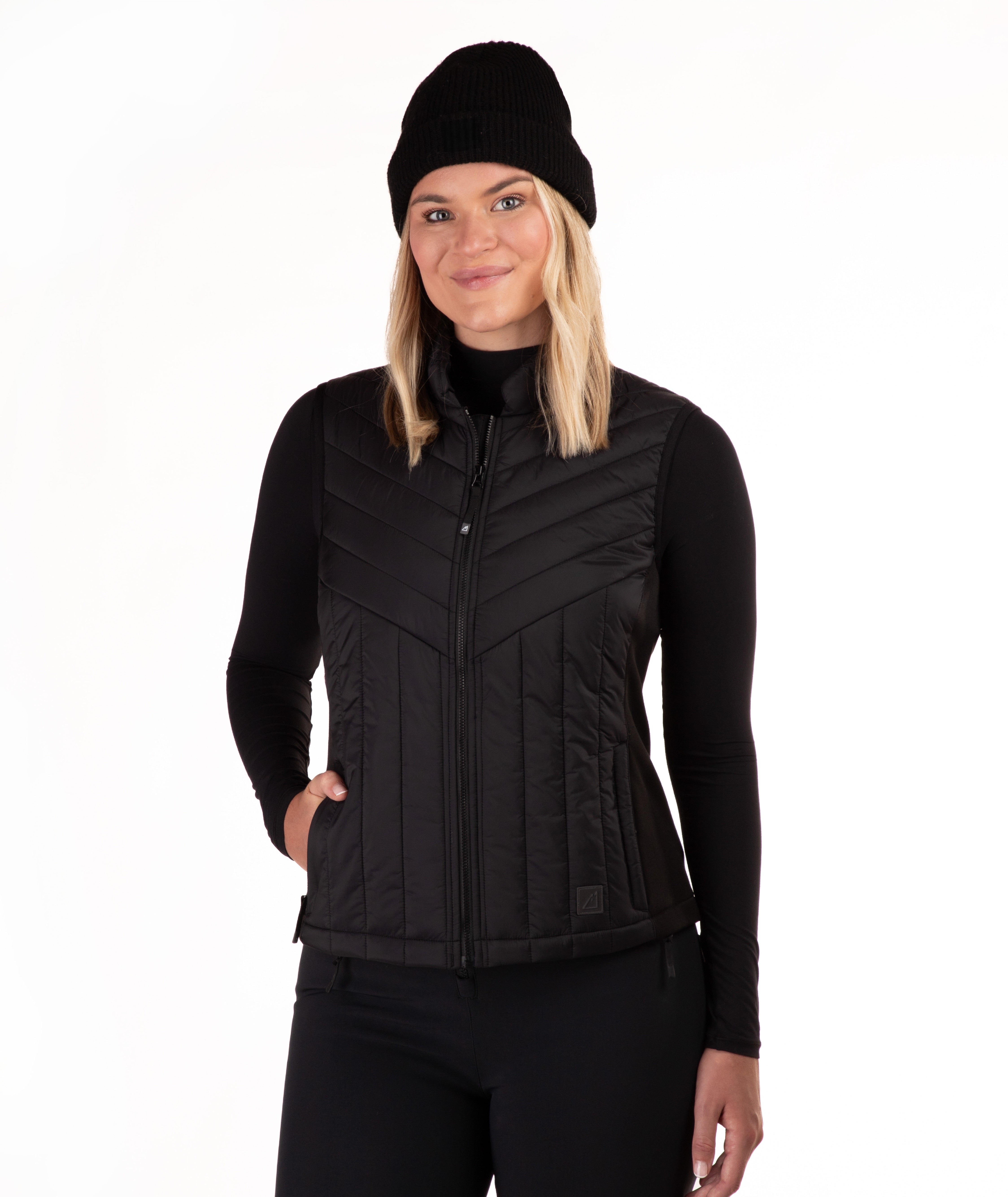A woman wearing a black zip-front quilted vest over a black long-sleeve top and black beanie, photographed against a plain white studio background.