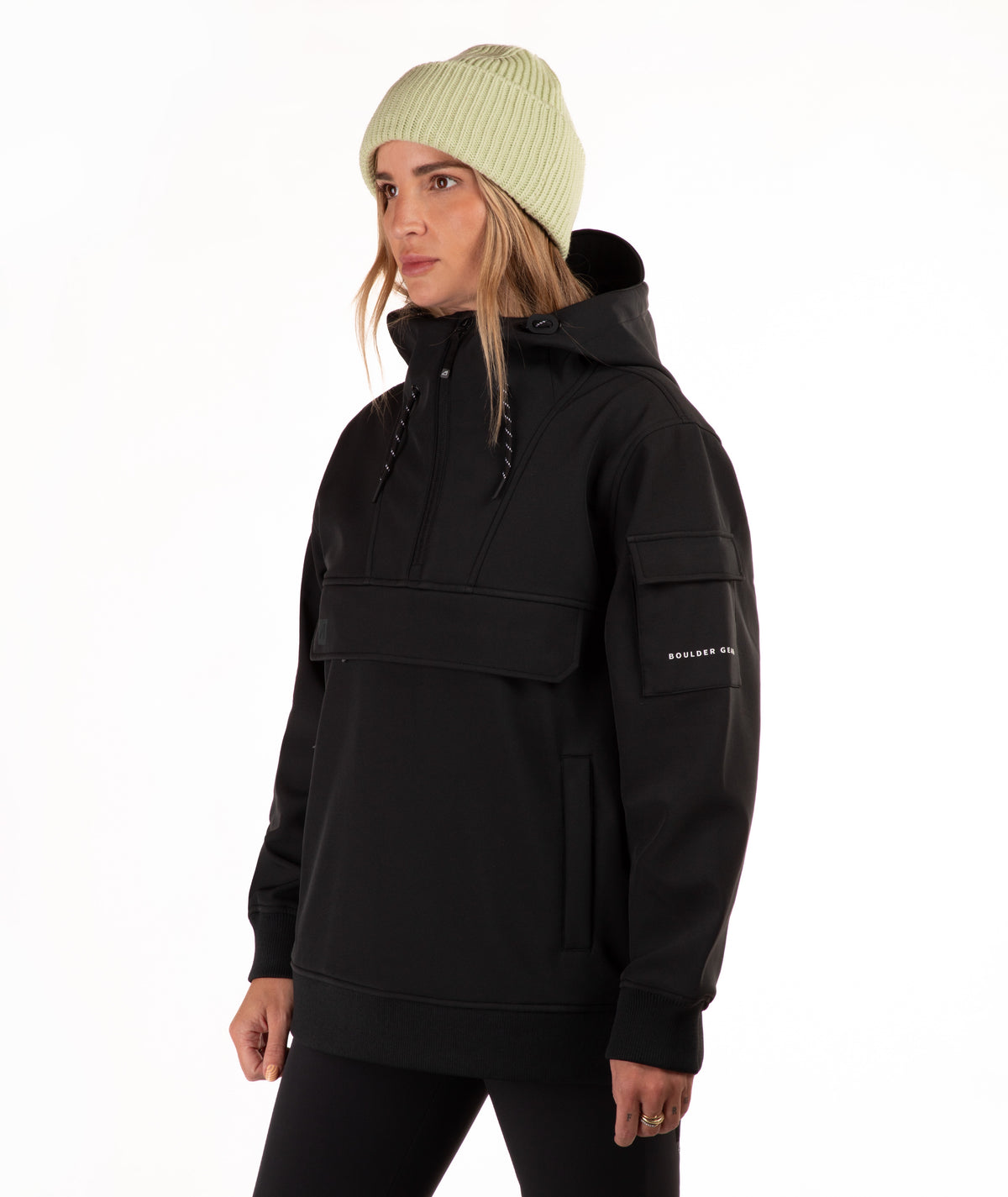 A woman wearing a black hooded pullover jacket with a high collar, drawstrings, and a horizontal chest pocket, paired with a light green ribbed knit beanie against a plain white background.