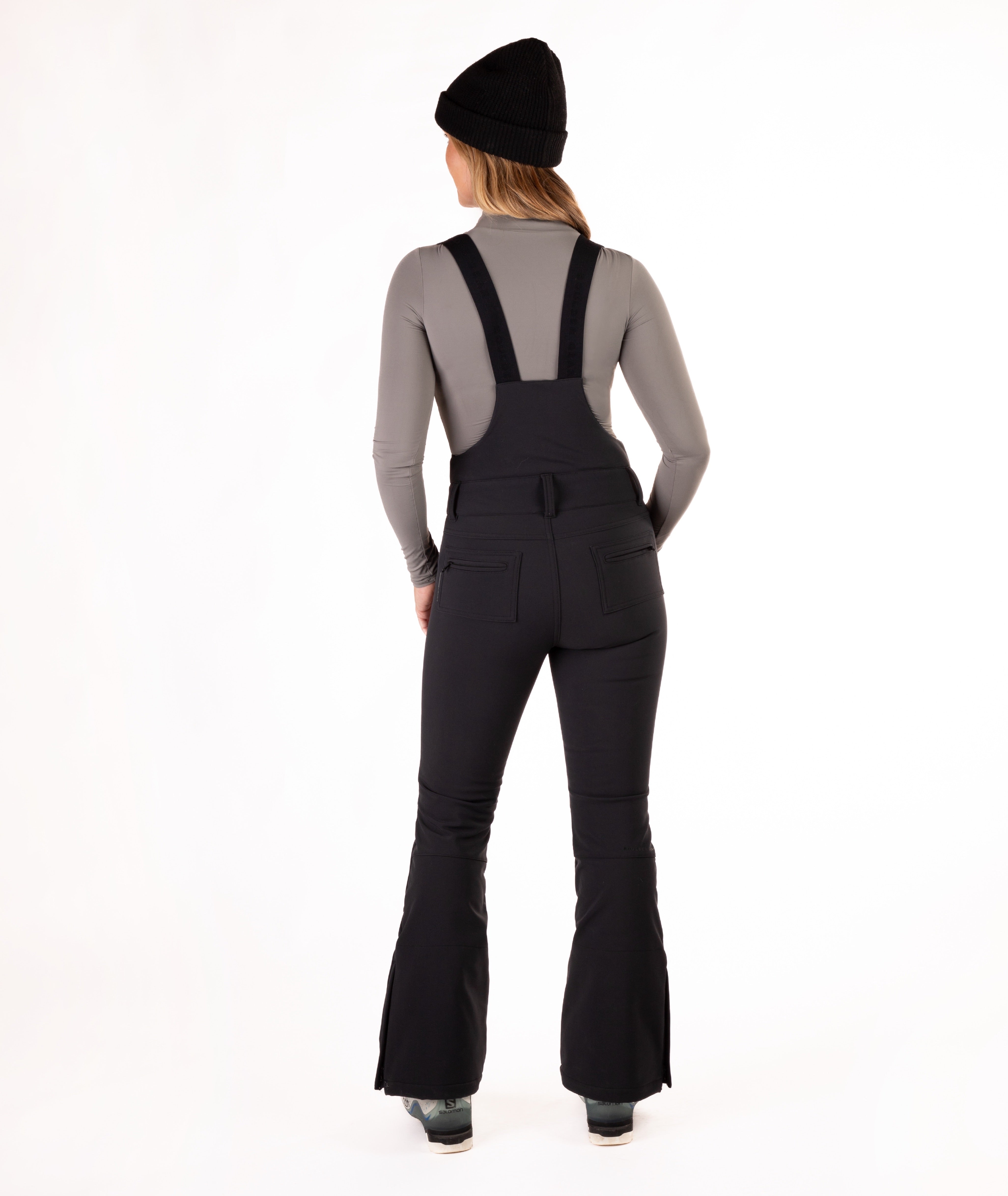 back of a Woman wearing black ski bib overalls with a gray top on a white background