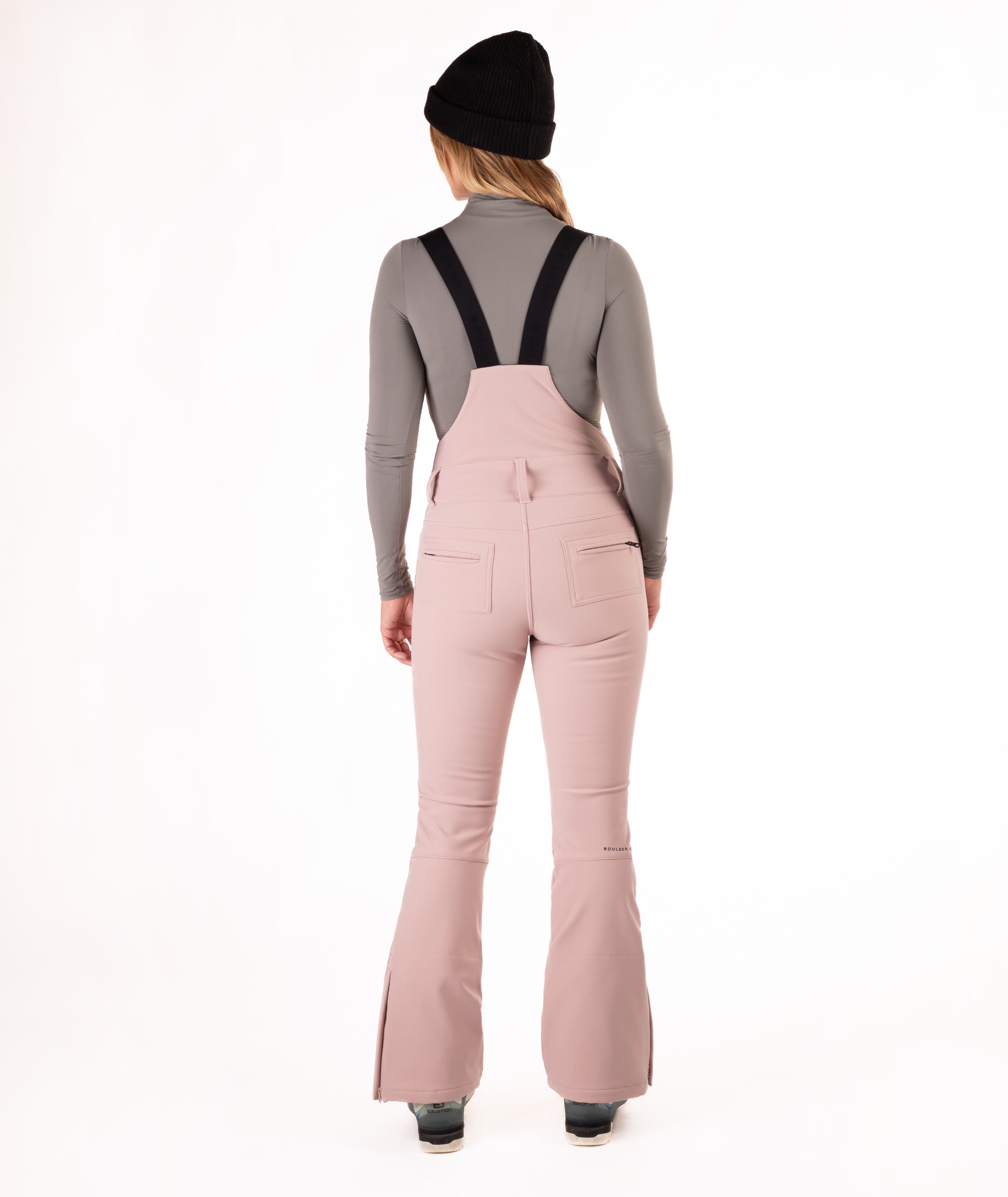 back of a Woman wearing pink ski bib overalls with a gray top on a white background