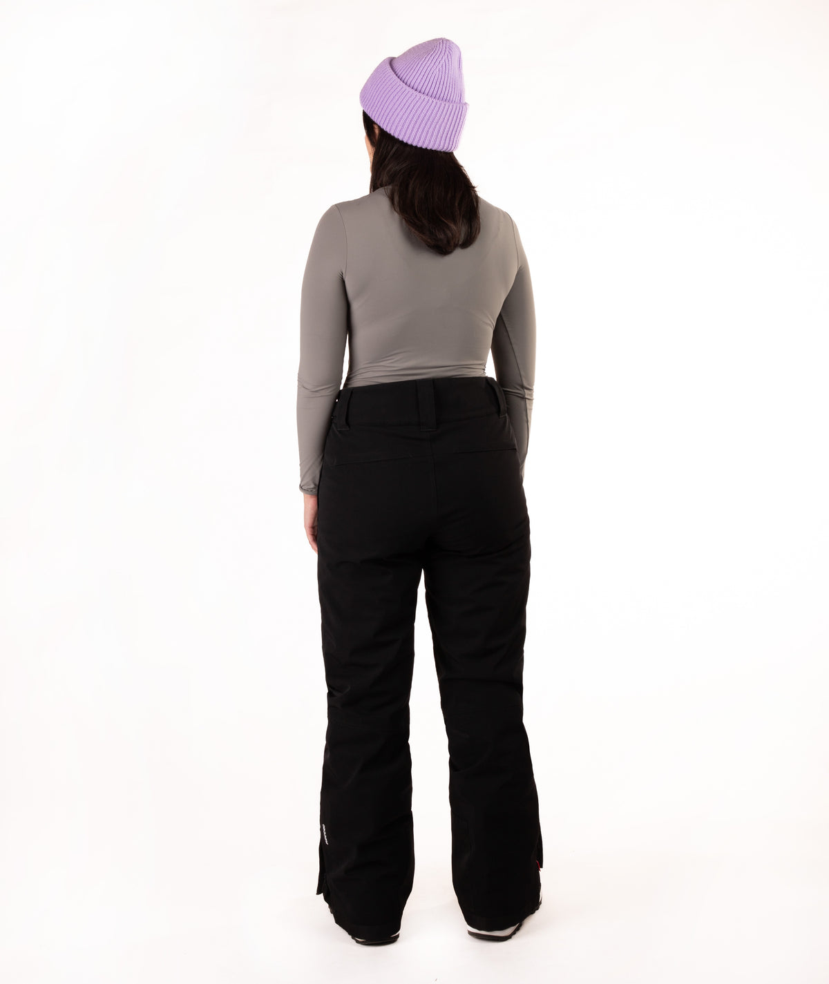A person shown from the back wearing a light purple knit beanie, a gray long-sleeve top, and black ski pants against a plain white studio background.