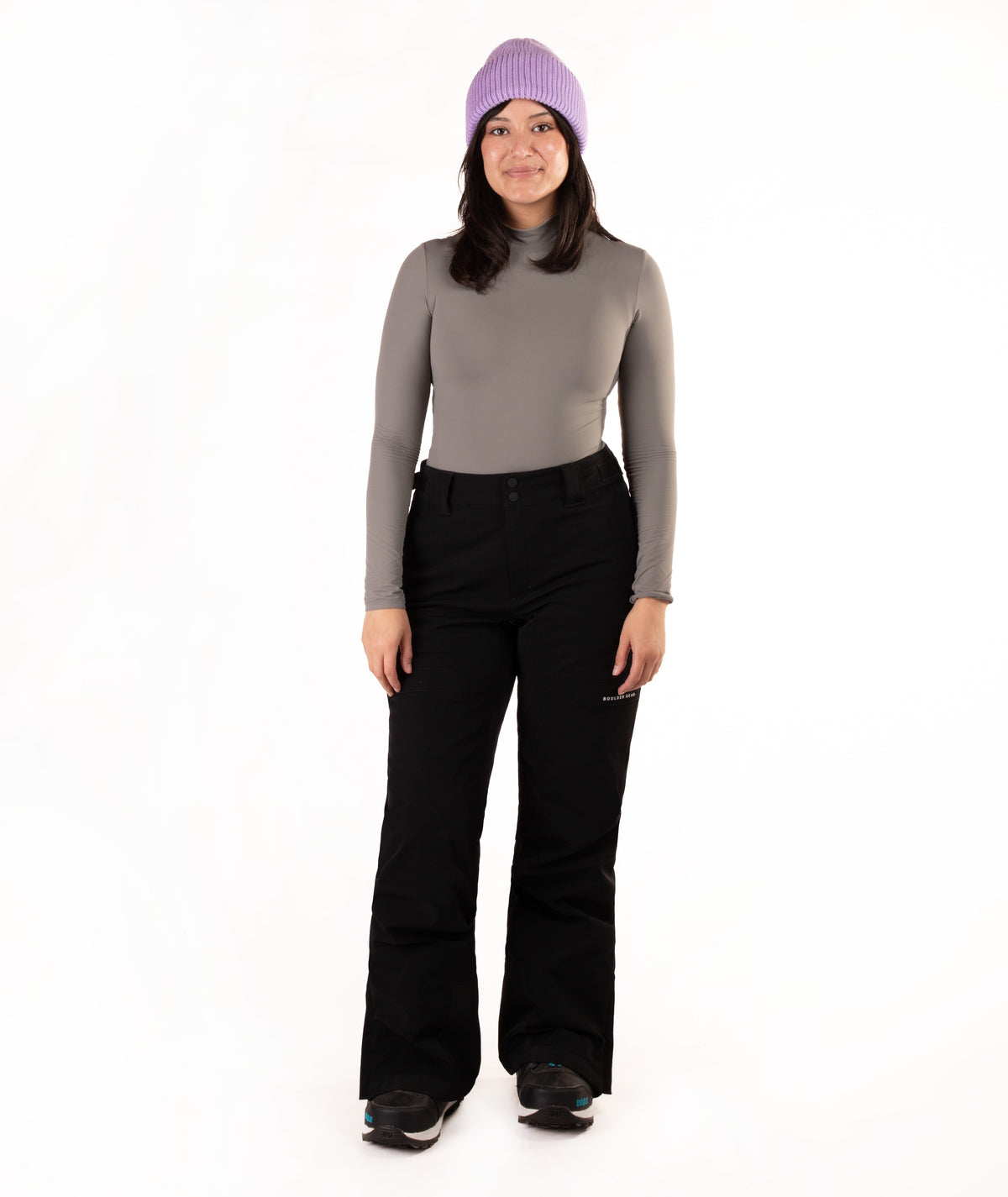 A woman modeling a gray long-sleeve mock neck top, black ski pants, and a lavender knit beanie against a plain white background.