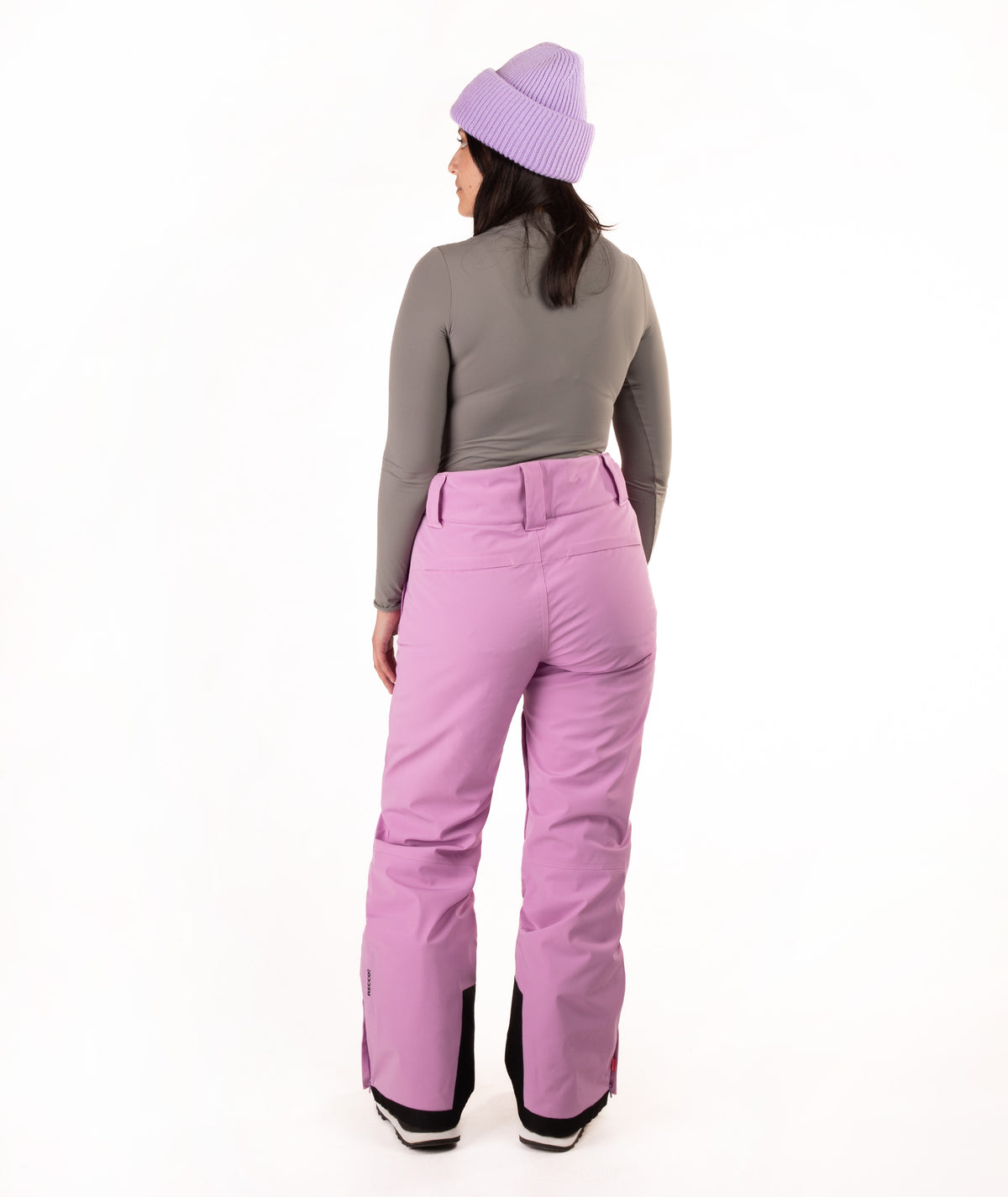 A woman viewed from the back wearing lavender snow pants, a gray long-sleeve top, and a light purple knit beanie against a plain white studio background.
