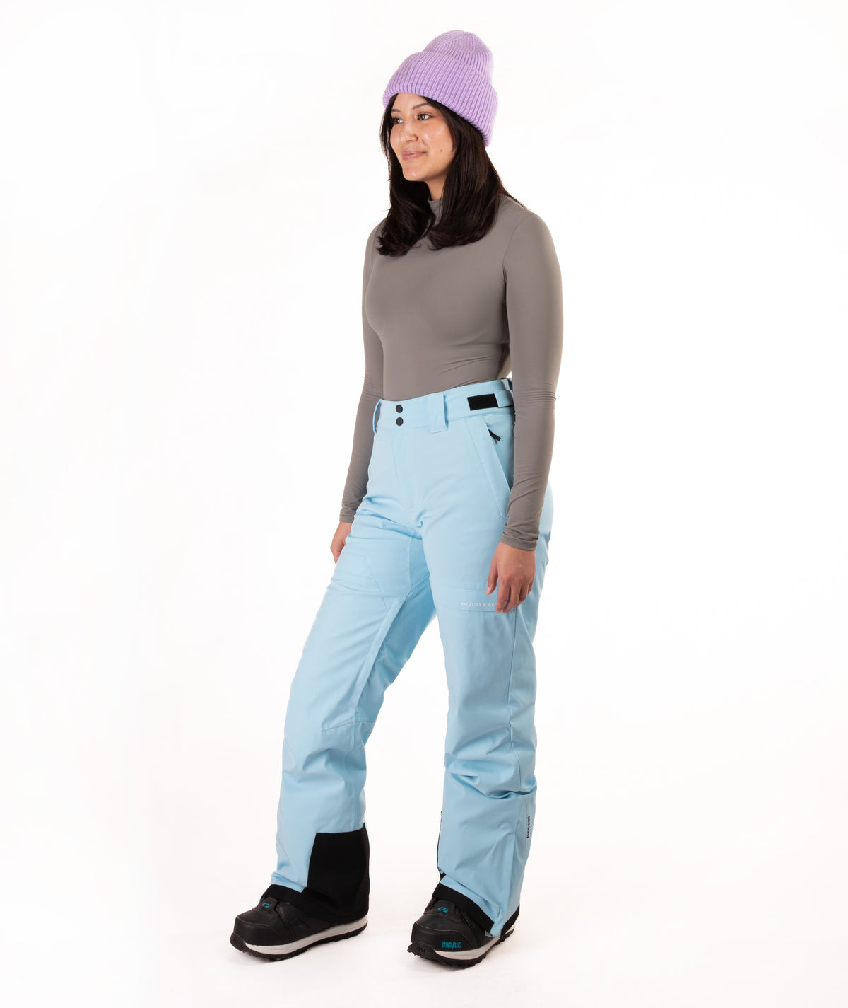 A woman modeling light blue snow pants, a gray long-sleeve top, a purple knit beanie, and black snow boots against a plain white studio background.