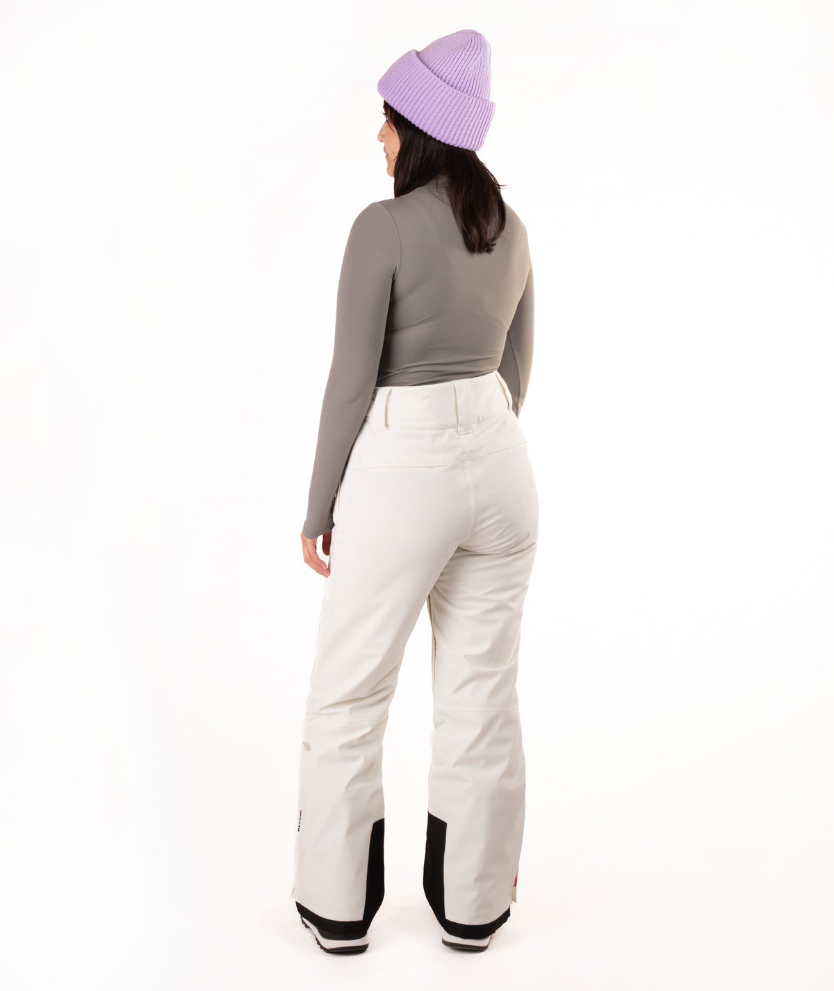 A woman shown from the back wearing a lavender knit beanie, a gray long-sleeve top, and off-white snow pants against a plain white studio background.