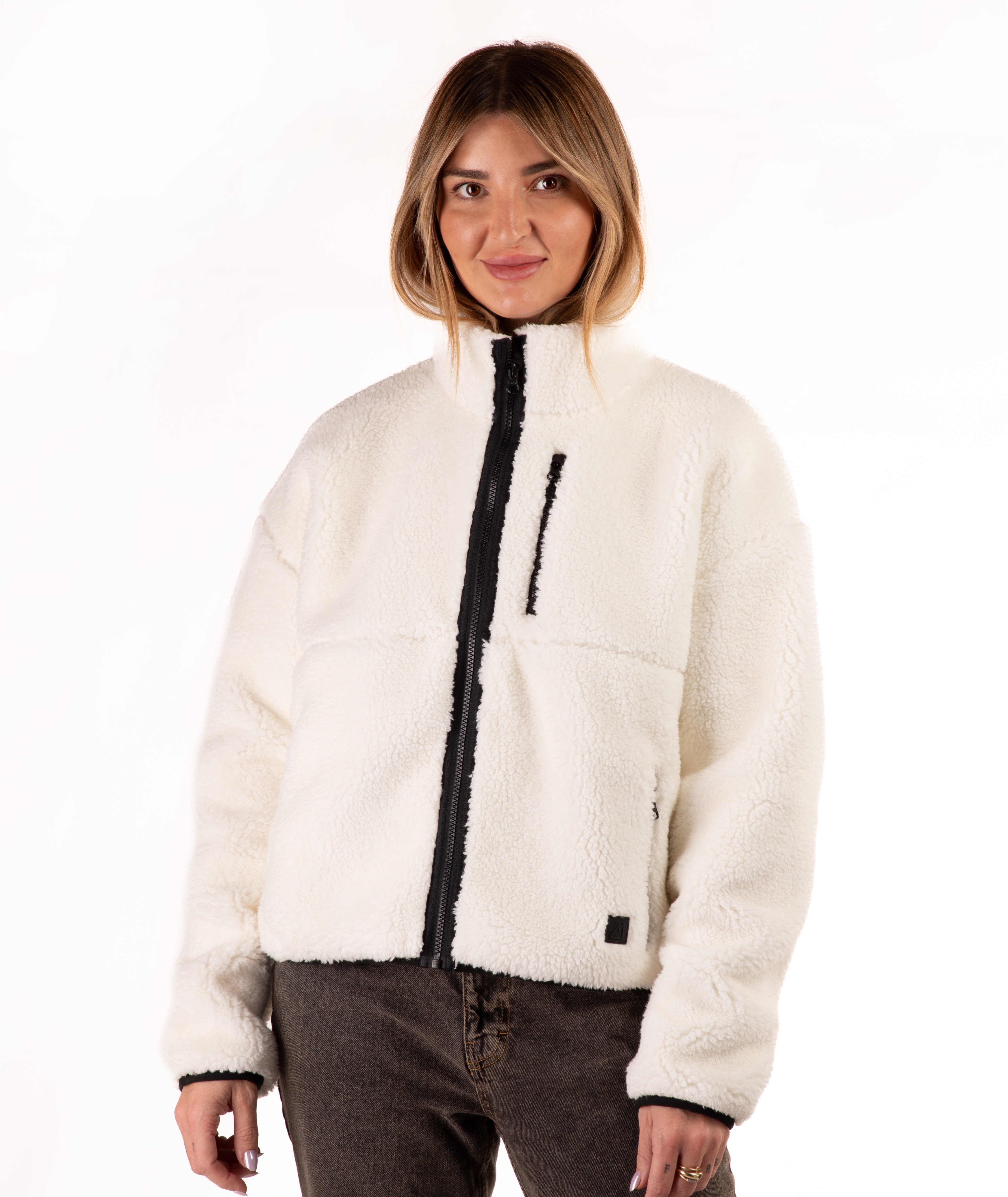 A woman wearing a off-white sherpa fleece zip-up jacket with black trim and a black front zipper, paired with dark brown jeans, against a plain white background.