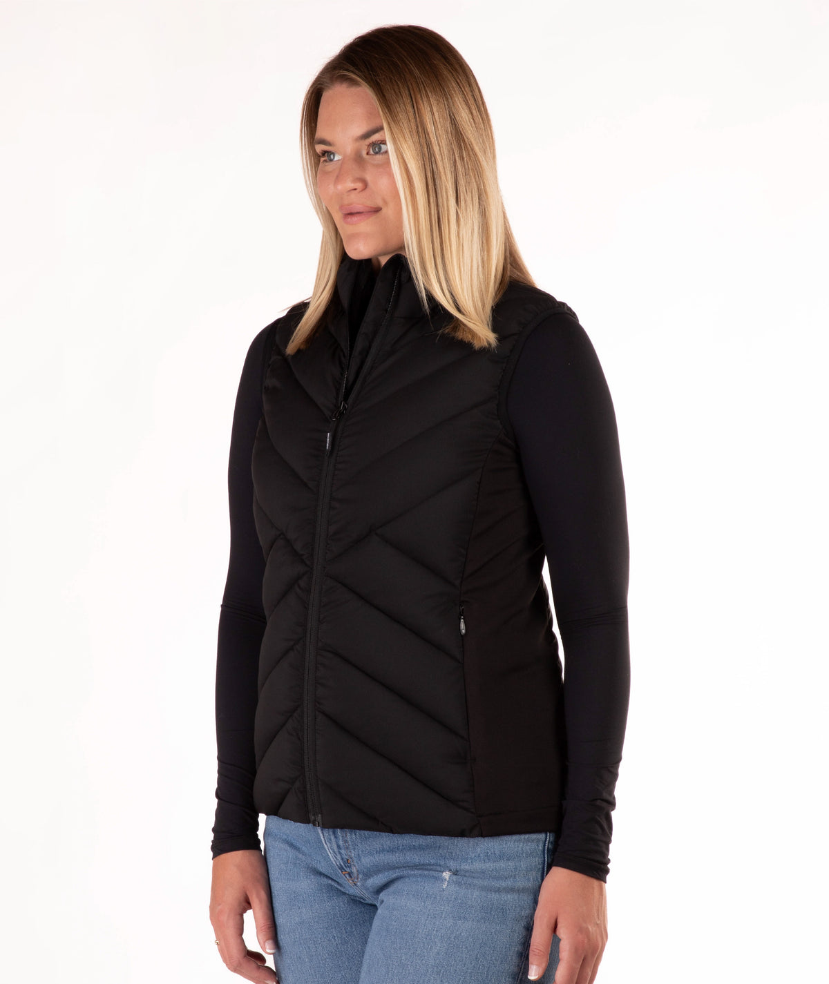 A woman modeling a black sleeveless zip-front puffer vest over a black long-sleeve top and blue jeans, photographed against a plain white studio background.