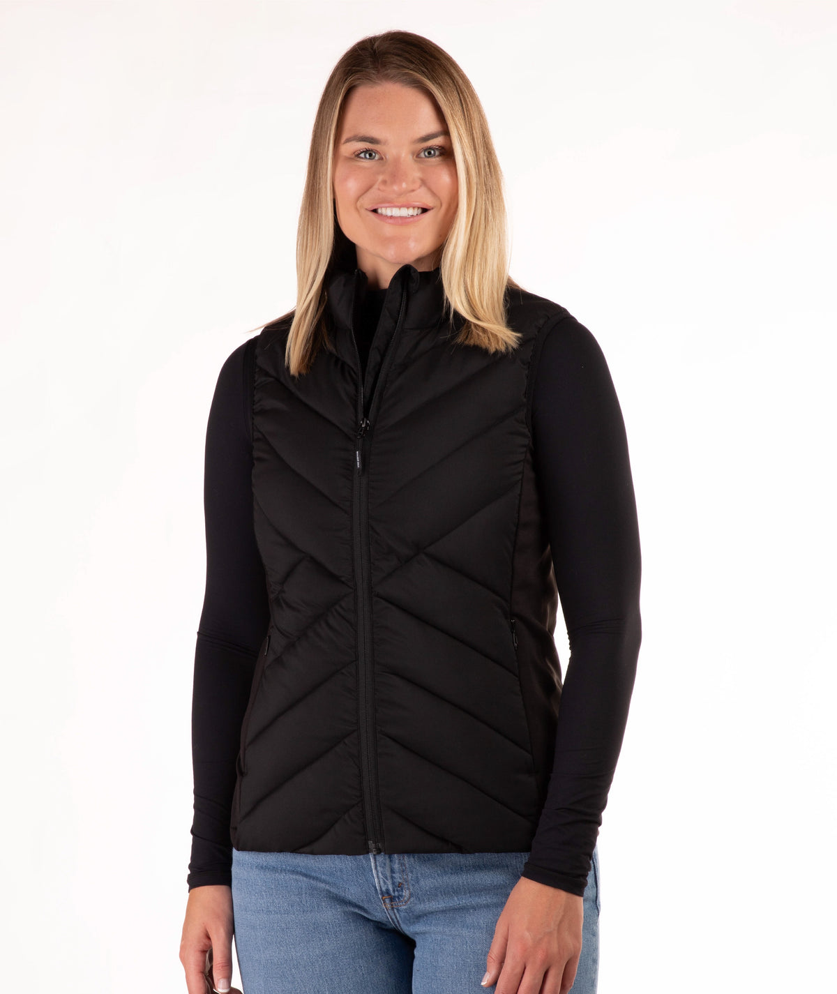 A woman modeling a black zip-front puffer vest over a black long-sleeve top and blue jeans, photographed against a plain white studio background.