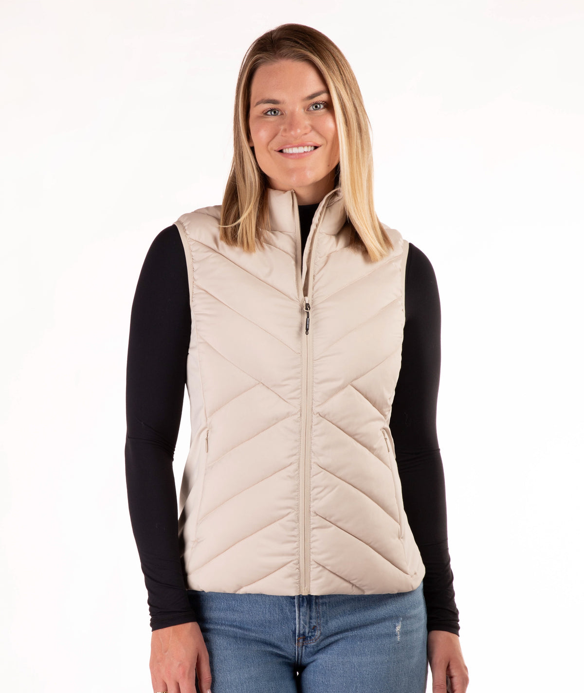 A woman modeling a light beige quilted puffer vest over a black long-sleeve shirt and blue jeans, photographed against a plain white studio background.
