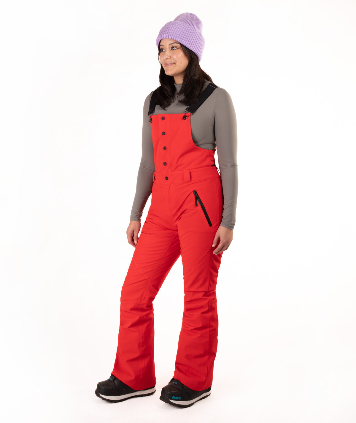 A woman modeling bright red snow bib overalls with black trim and buttons, worn over a gray long-sleeve top, black boots, and a lavender knit beanie against a plain white background.