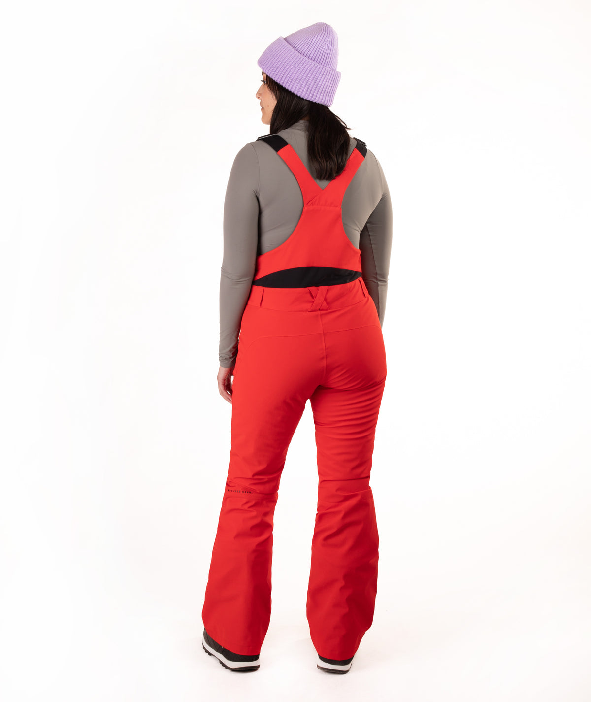 A woman shown from the back wearing bright red ski bib pants over a gray long-sleeve top, with a light purple knit beanie, against a plain white studio background.