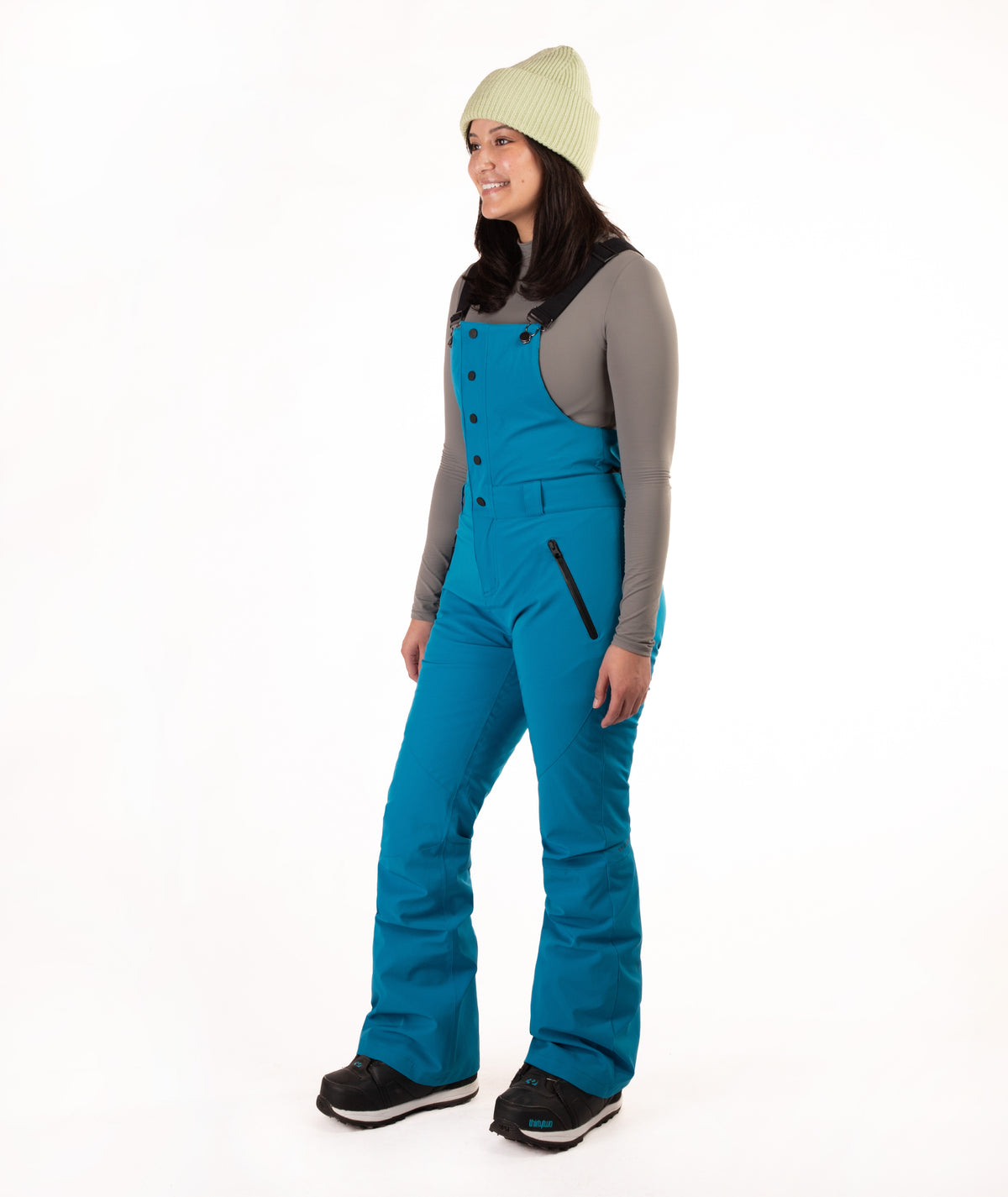 A woman modeling bright teal snow bib overalls with a gray long-sleeve shirt, a light green knit beanie, and black boots, standing against a solid white studio background.