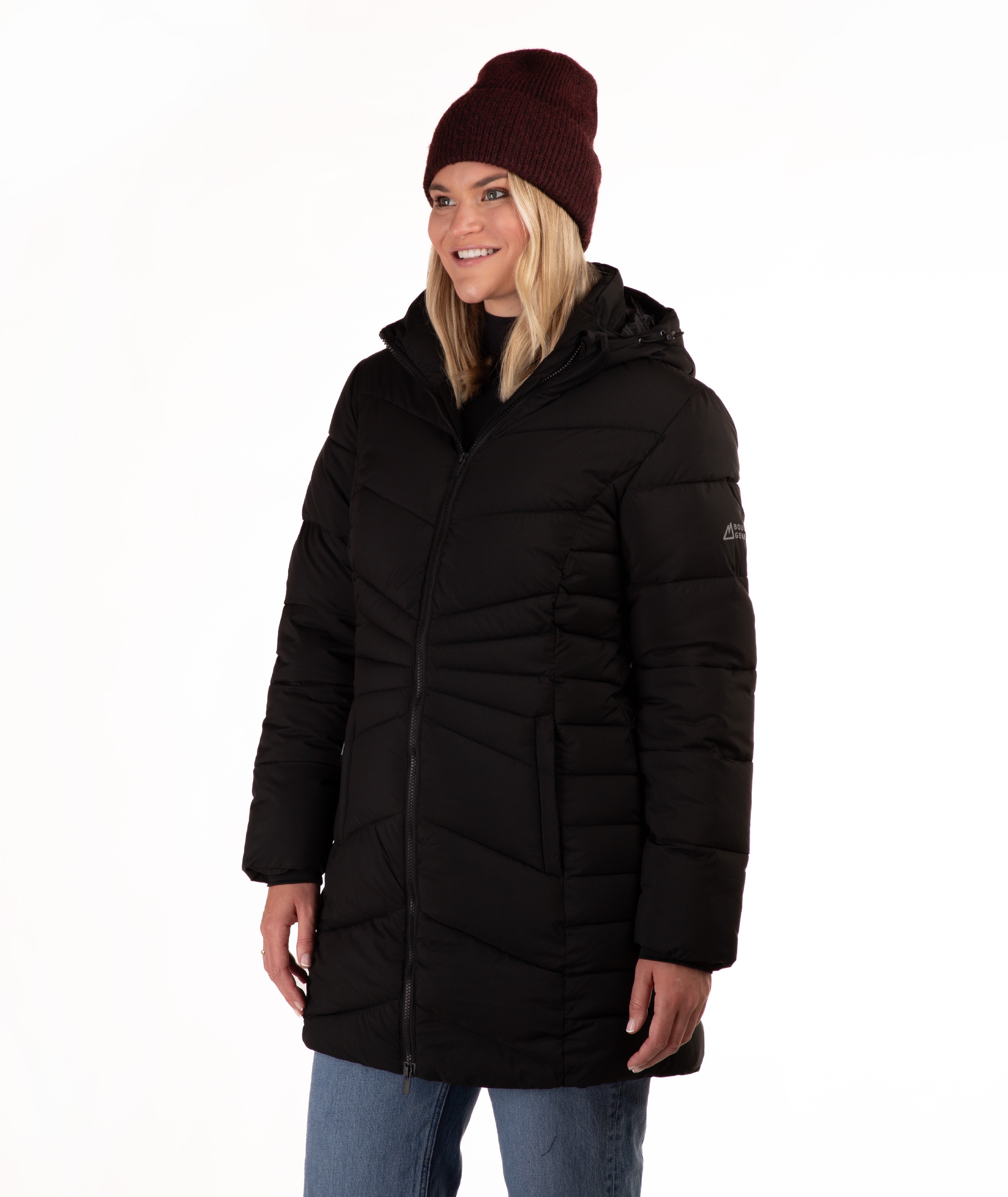 A woman wearing a black hooded puffer jacket, a dark burgundy knit beanie, and blue jeans, photographed against a plain white studio background.