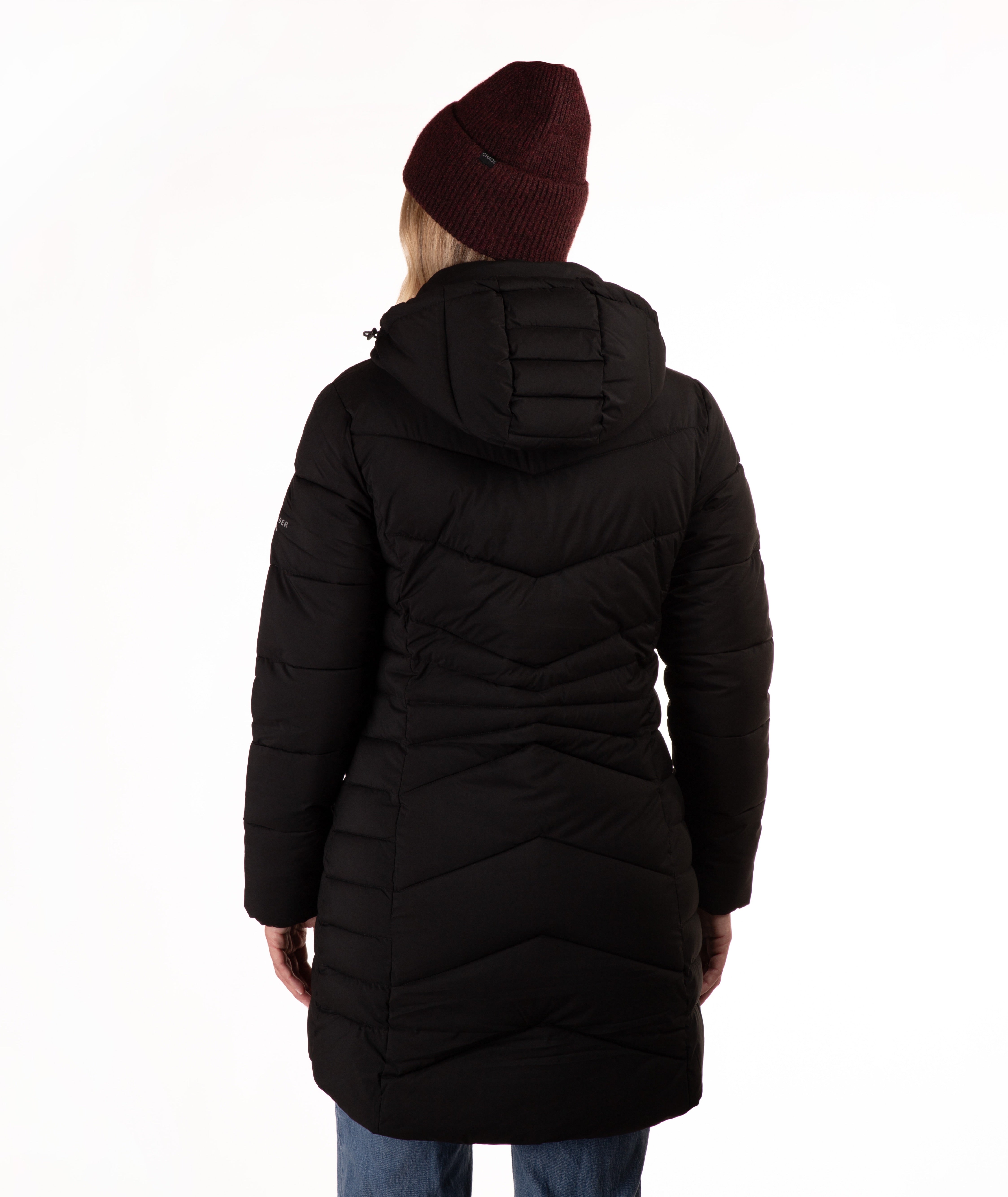 A woman viewed from behind wearing a long black hooded puffer coat, a dark burgundy knit beanie, and blue jeans against a plain white studio background.