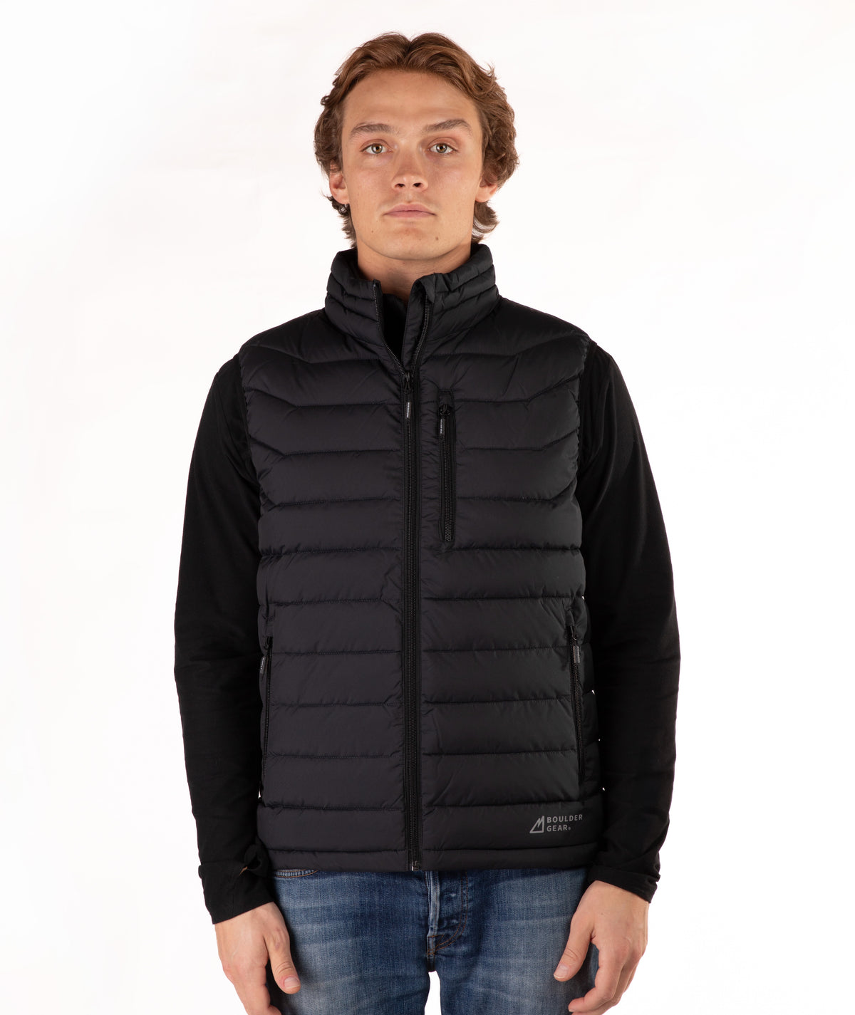 A man wearing a black quilted zip-up puffer vest over a black long-sleeve shirt and blue jeans, standing against a plain white studio background.