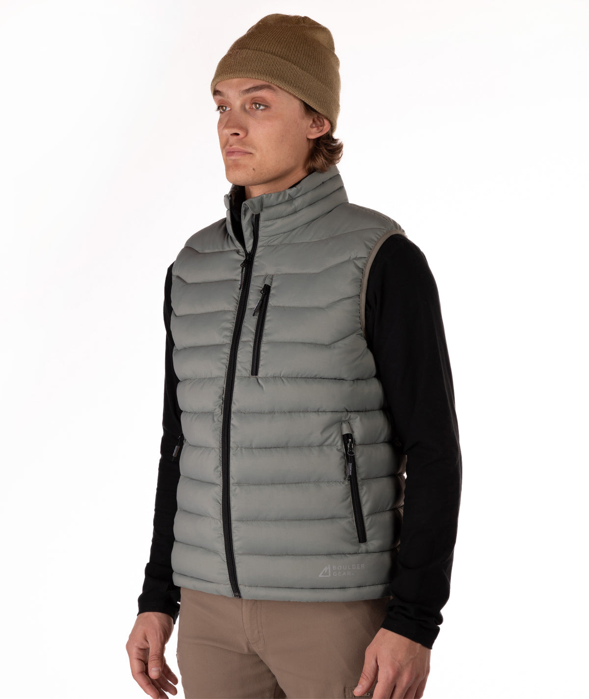 A man wearing a taupe puffer vest over a black long-sleeve shirt, with a brown beanie and khaki pants, standing against a plain white studio background.