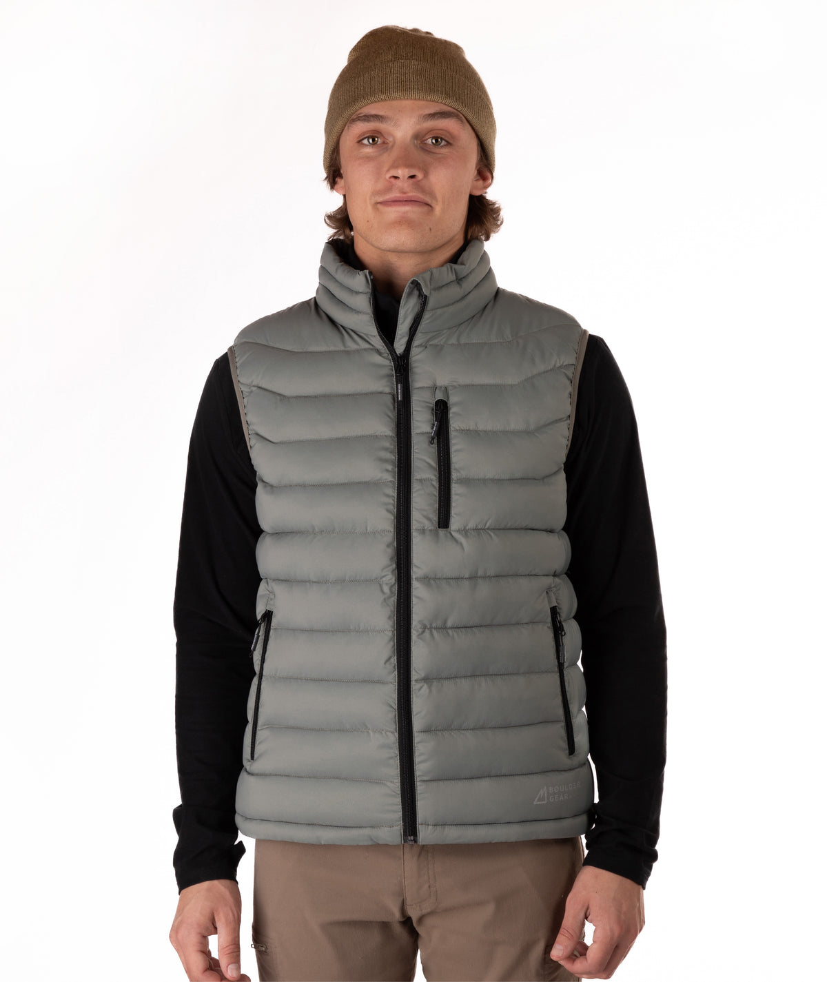 A man wearing a taupe puffer vest over a black long-sleeve shirt, with a brown beanie and khaki pants, standing against a plain white studio background.