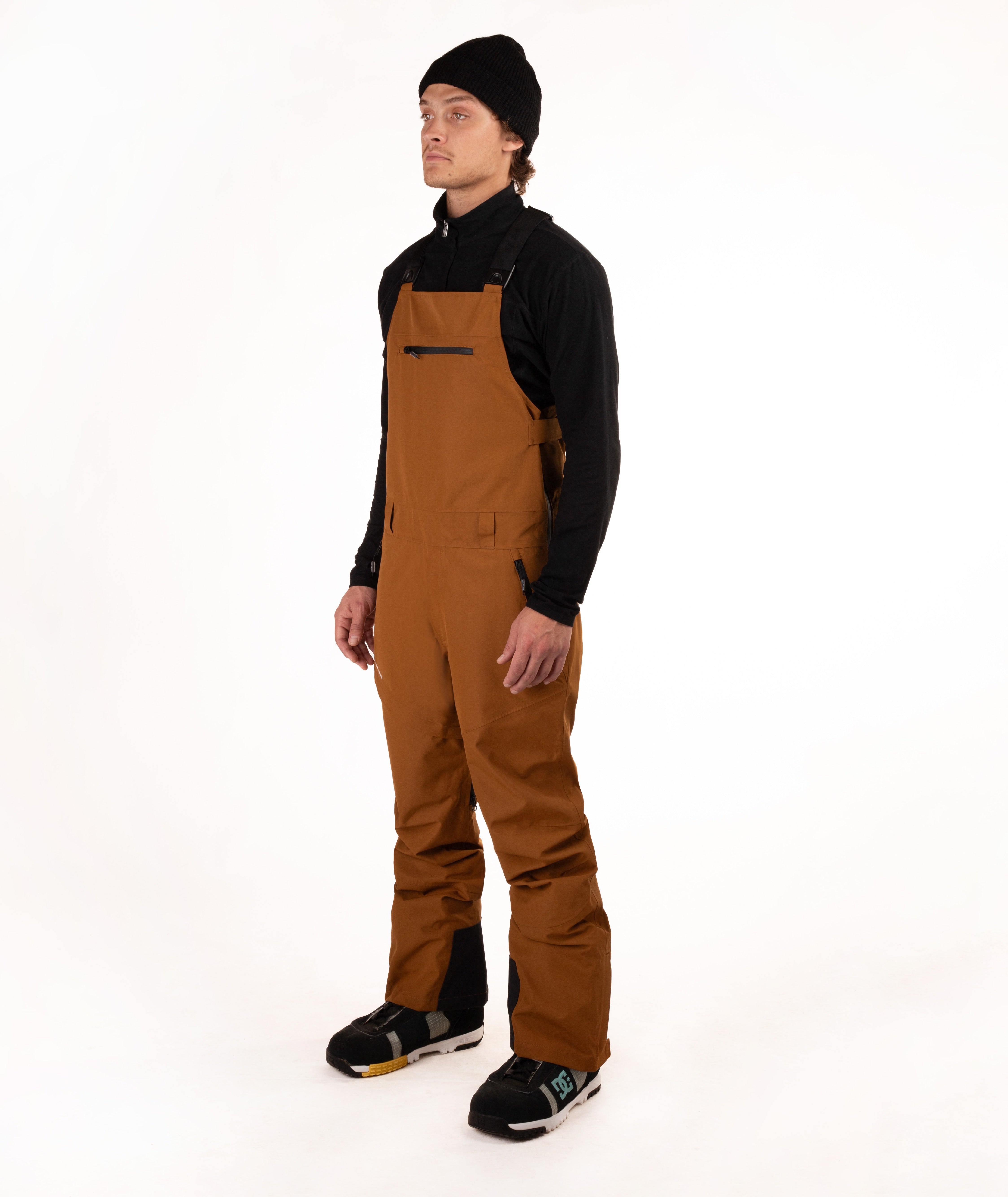 A male model wearing brown snow bib overalls with black panels, a black long-sleeve shirt, and a black beanie, standing against a solid white studio background.