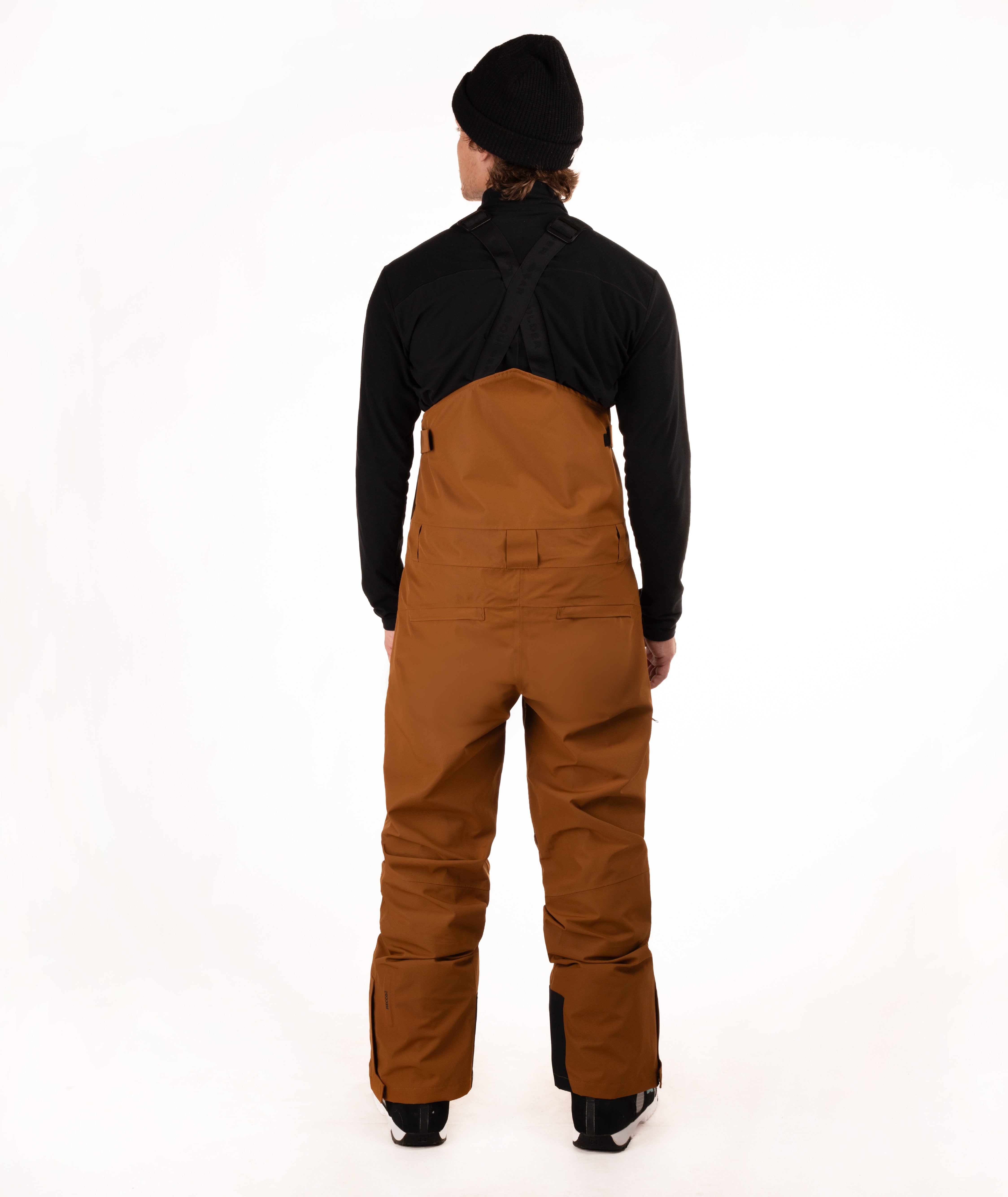 A model shown from the back wearing brown snow bib overalls over a black long-sleeve top and black beanie, standing against a white studio background.