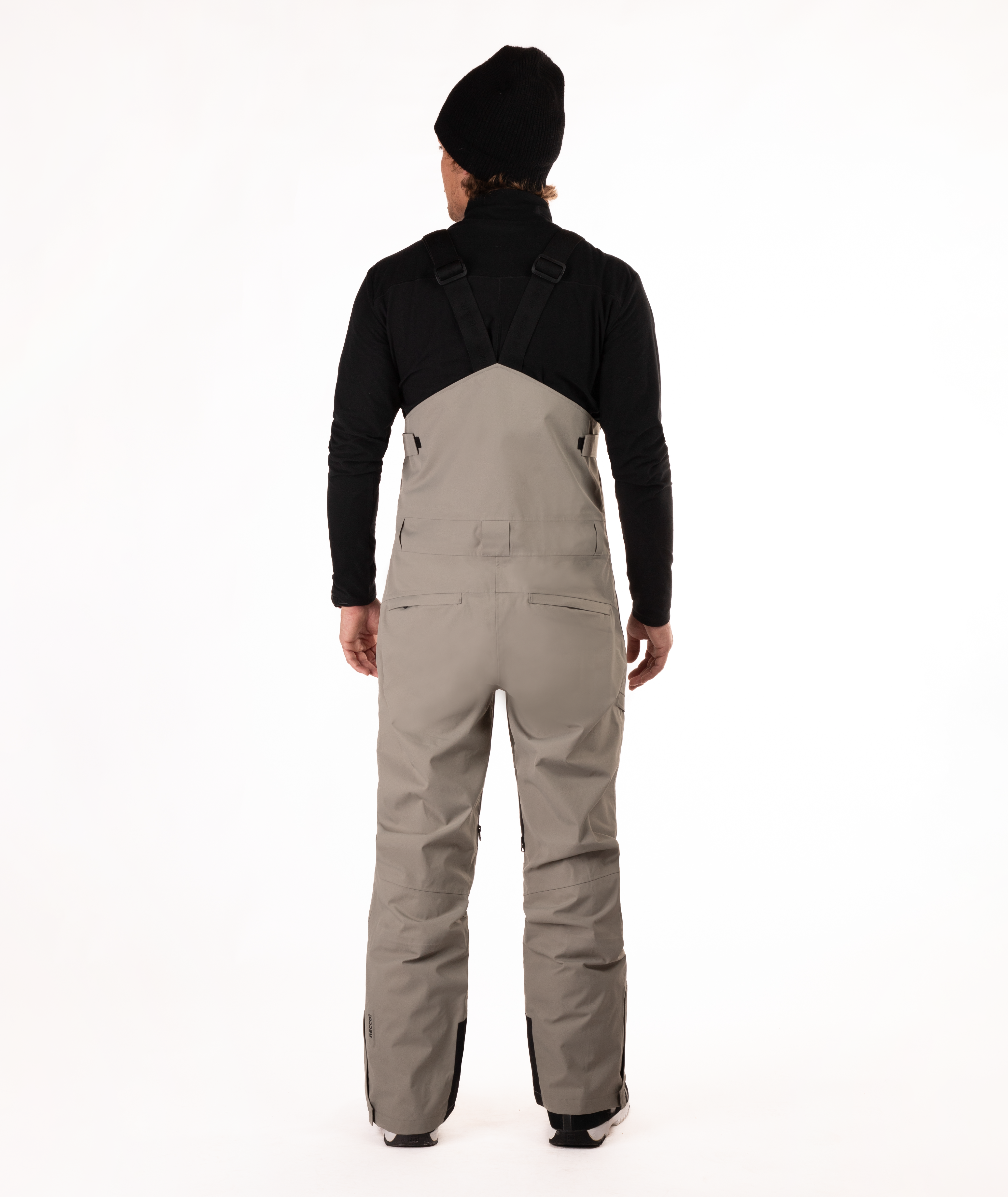 back of Person wearing a black long-sleeve shirt and beige bib overalls on a white background