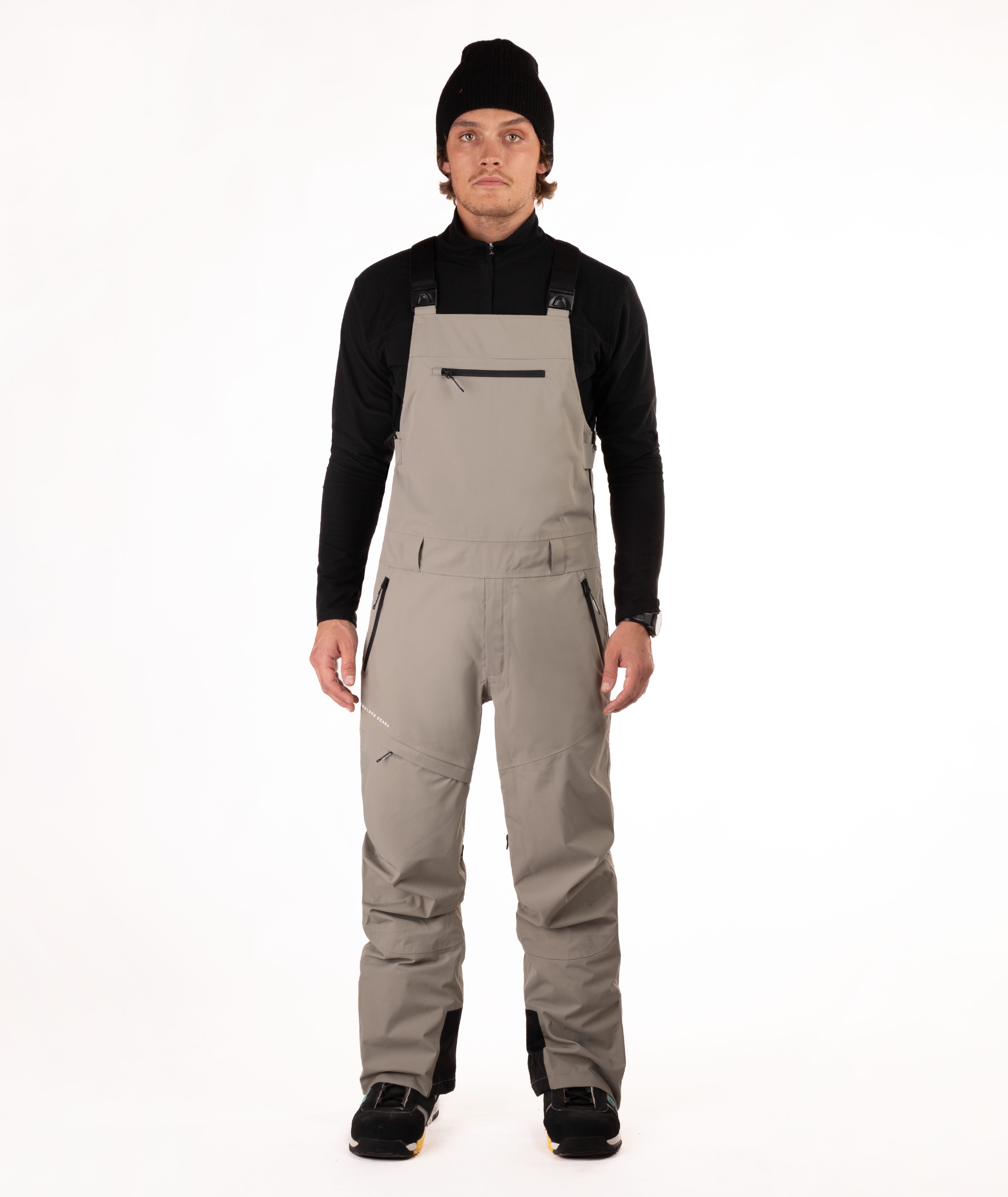 Person wearing a black long-sleeve shirt and beige bib overalls on a white background