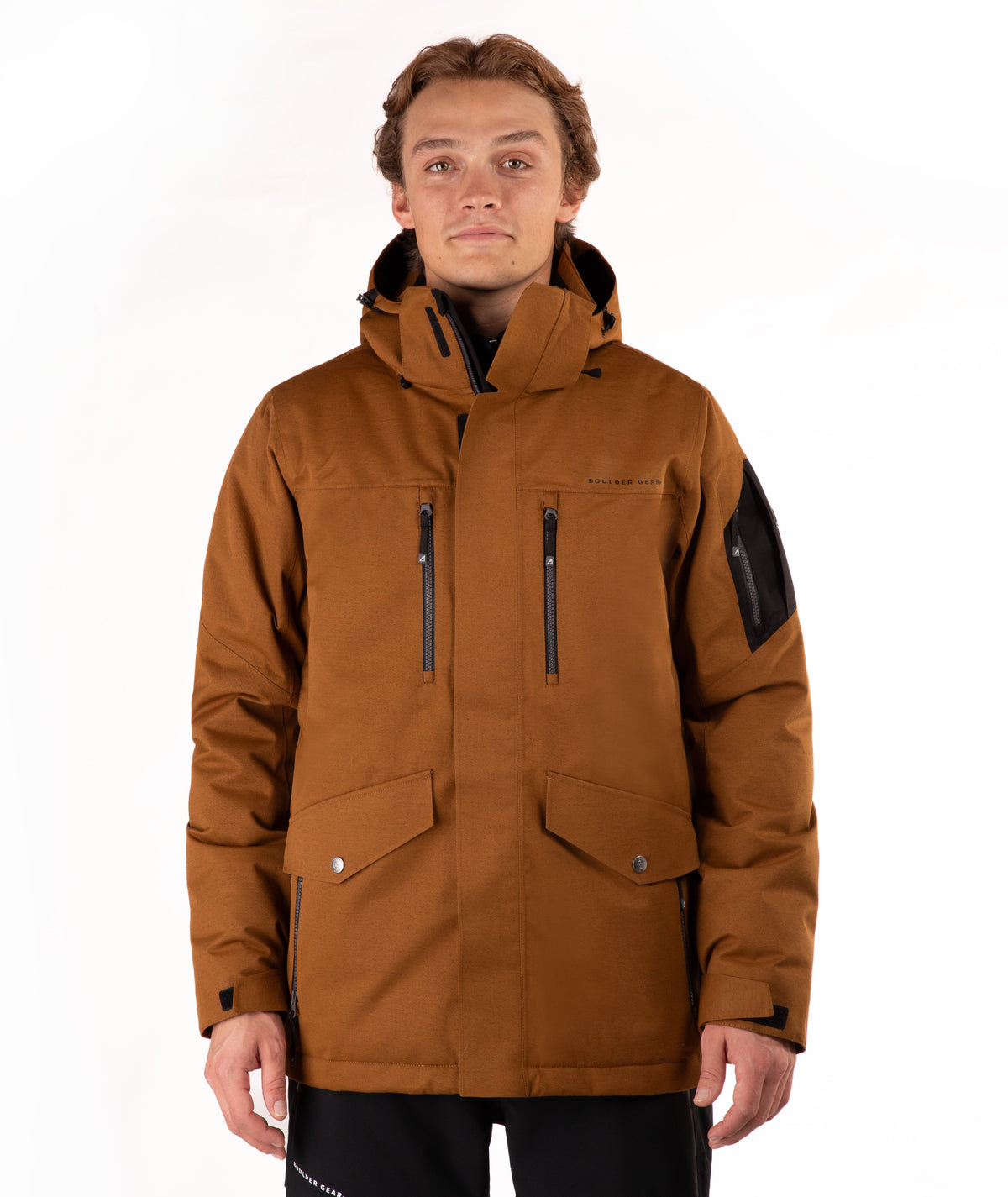A man wearing a brown hooded technical jacket with black zippers, flap pockets, and a black sleeve patch, photographed against a plain white studio background.