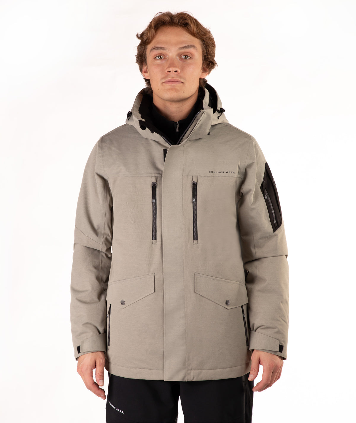 A man wearing a taupe hooded technical jacket with black zippers, black sleeve panels, and a black collar, standing against a plain white background.