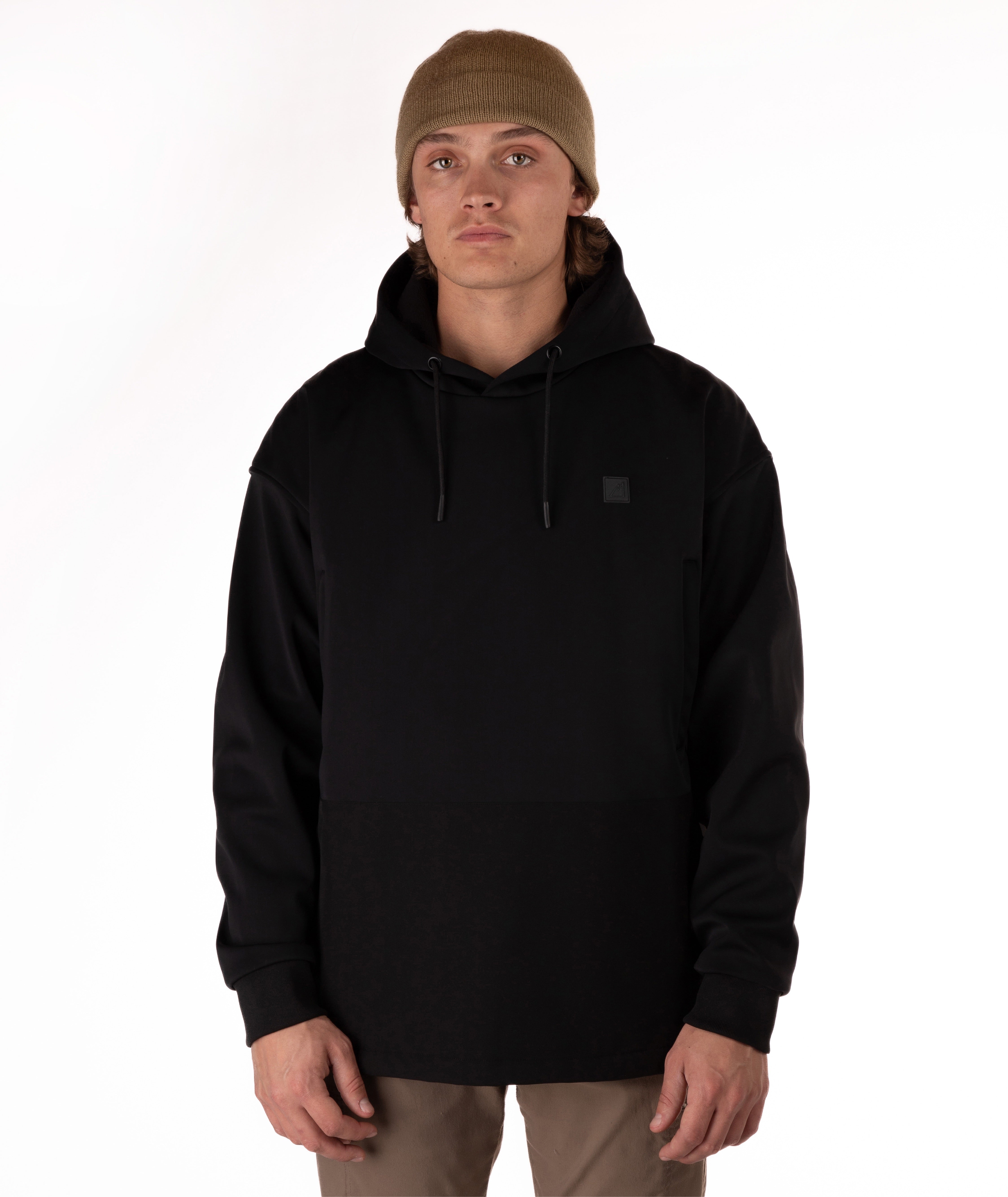 A man wearing a black pullover hoodie with a small square logo patch on the left chest, paired with a brown beanie and khaki pants, photographed against a plain white studio background.