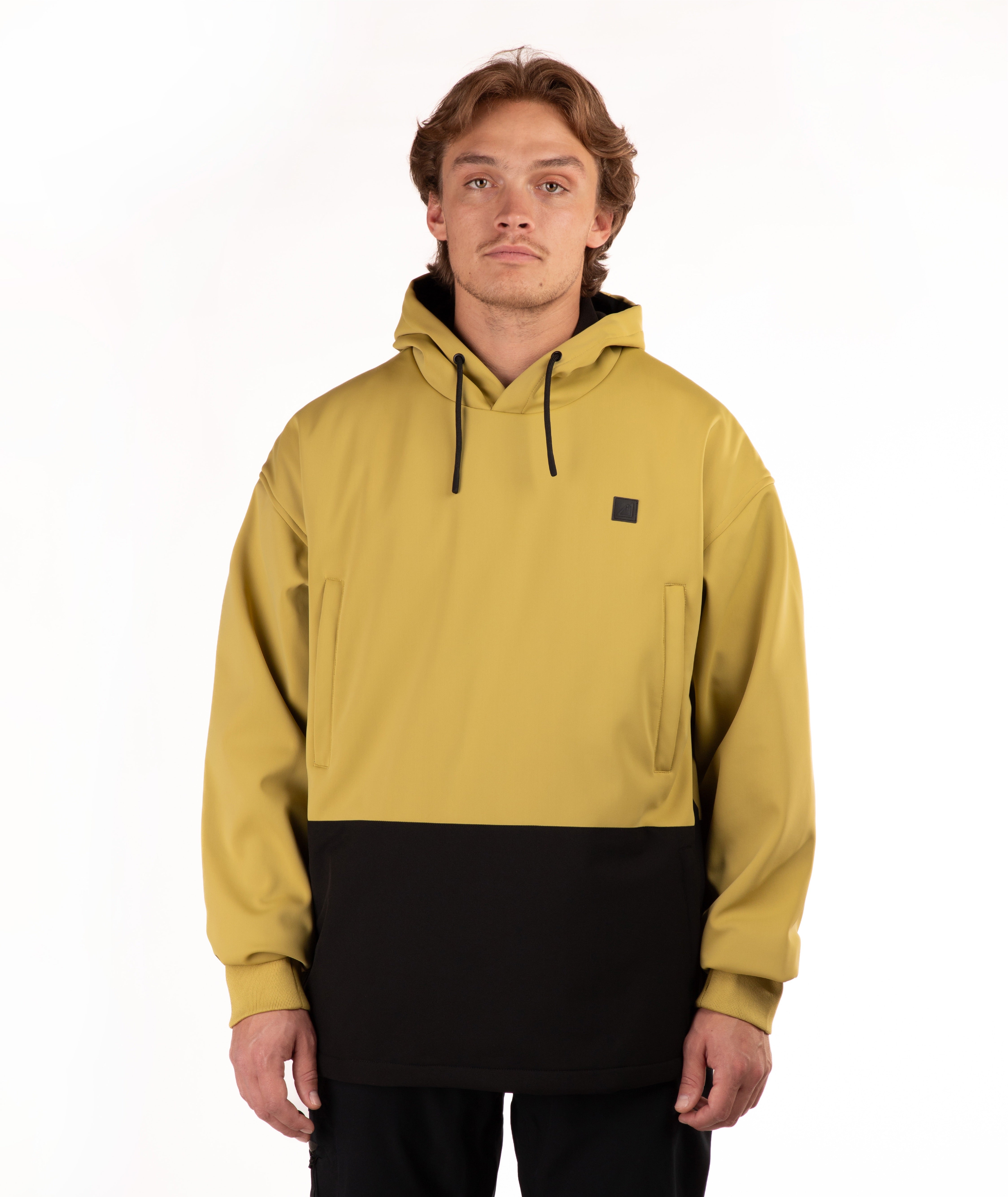 A man wearing a color-block pullover hoodie in mustard yellow and black, featuring a small black square logo on the left chest, photographed against a plain white studio background.