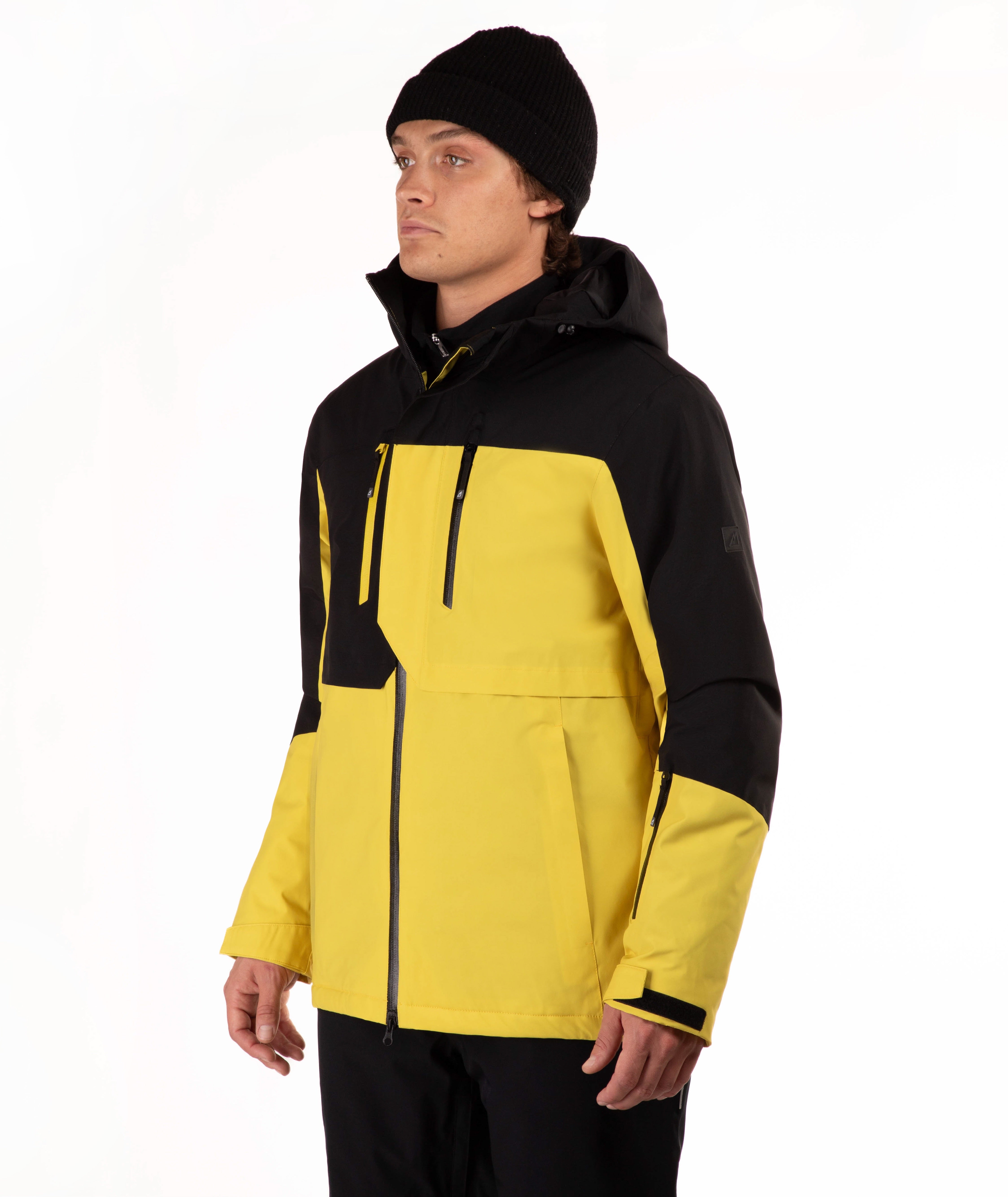 A man wearing a yellow and black ski jacket, black pants, and a black beanie, standing against a plain white studio background.