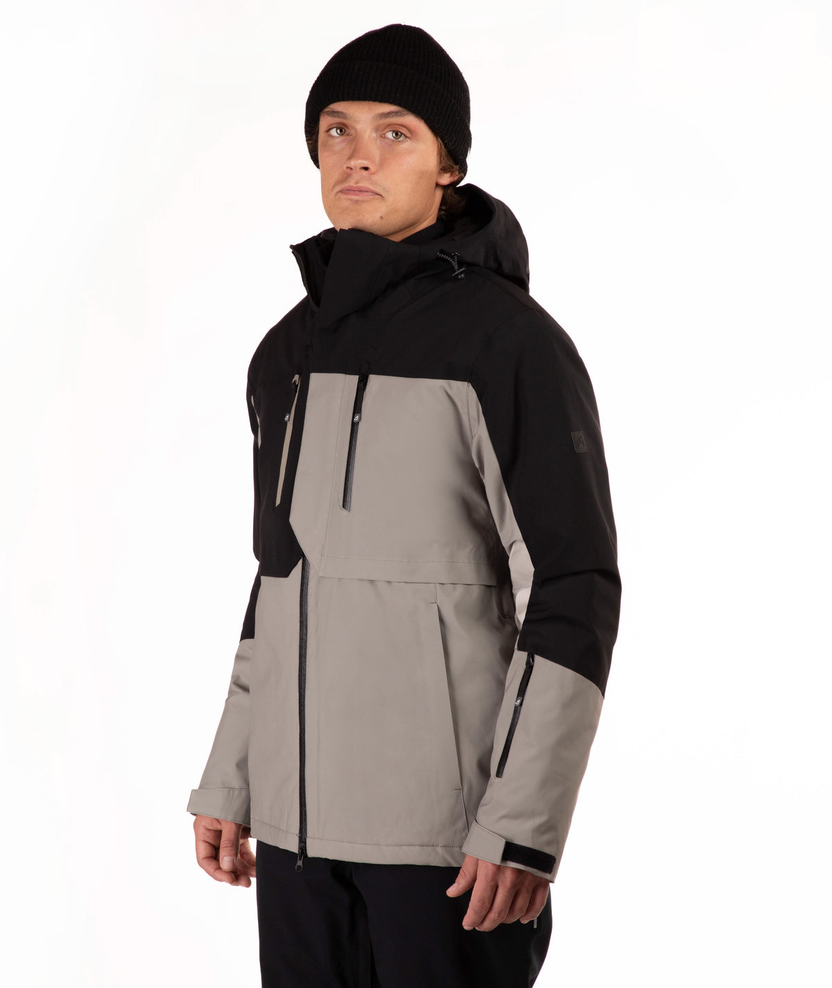 A man wearing a color-blocked ski jacket in black and taupe, paired with a black beanie, photographed against a plain white studio background.