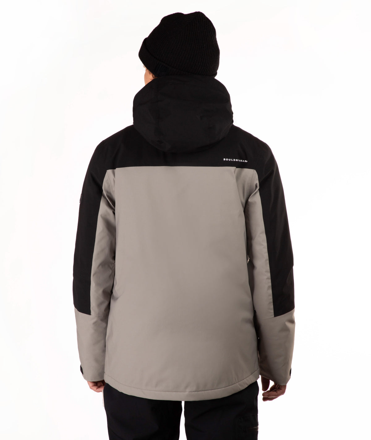 A person shown from the back wearing a two-tone outdoor jacket with a black hood and black sleeves, paired with a taupe body panel, black pants, and a black beanie against a plain white background.