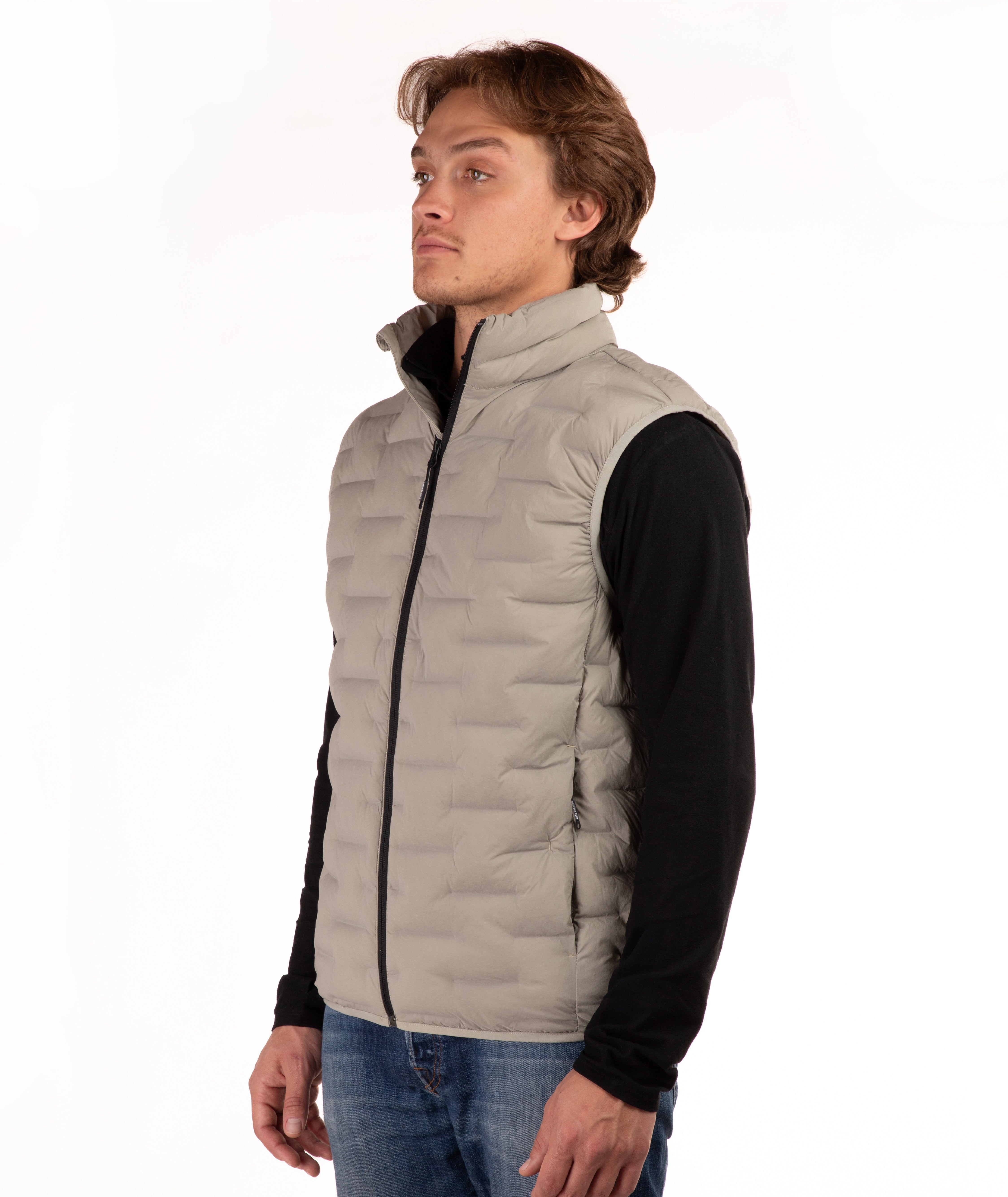 A man wearing a light beige quilted puffer vest over a black long-sleeve shirt and blue jeans, standing against a plain white background.