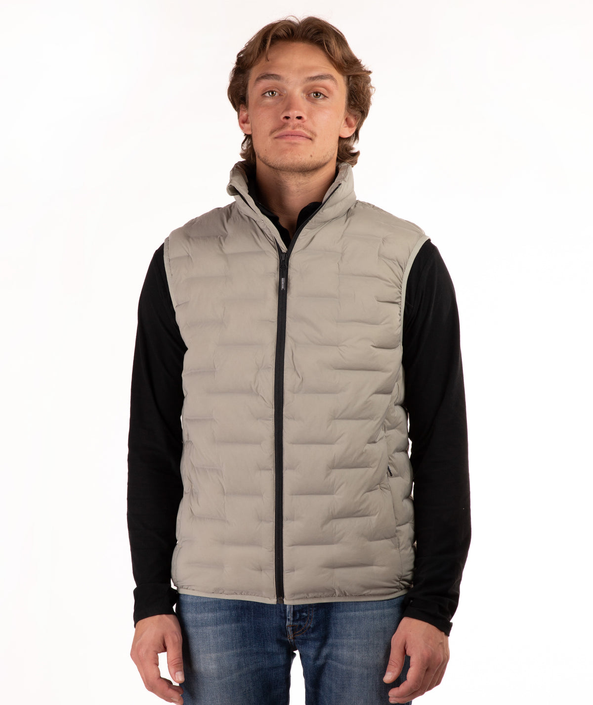A man wearing a light beige quilted puffer vest over a black long-sleeve shirt and blue jeans, standing against a plain white background.
