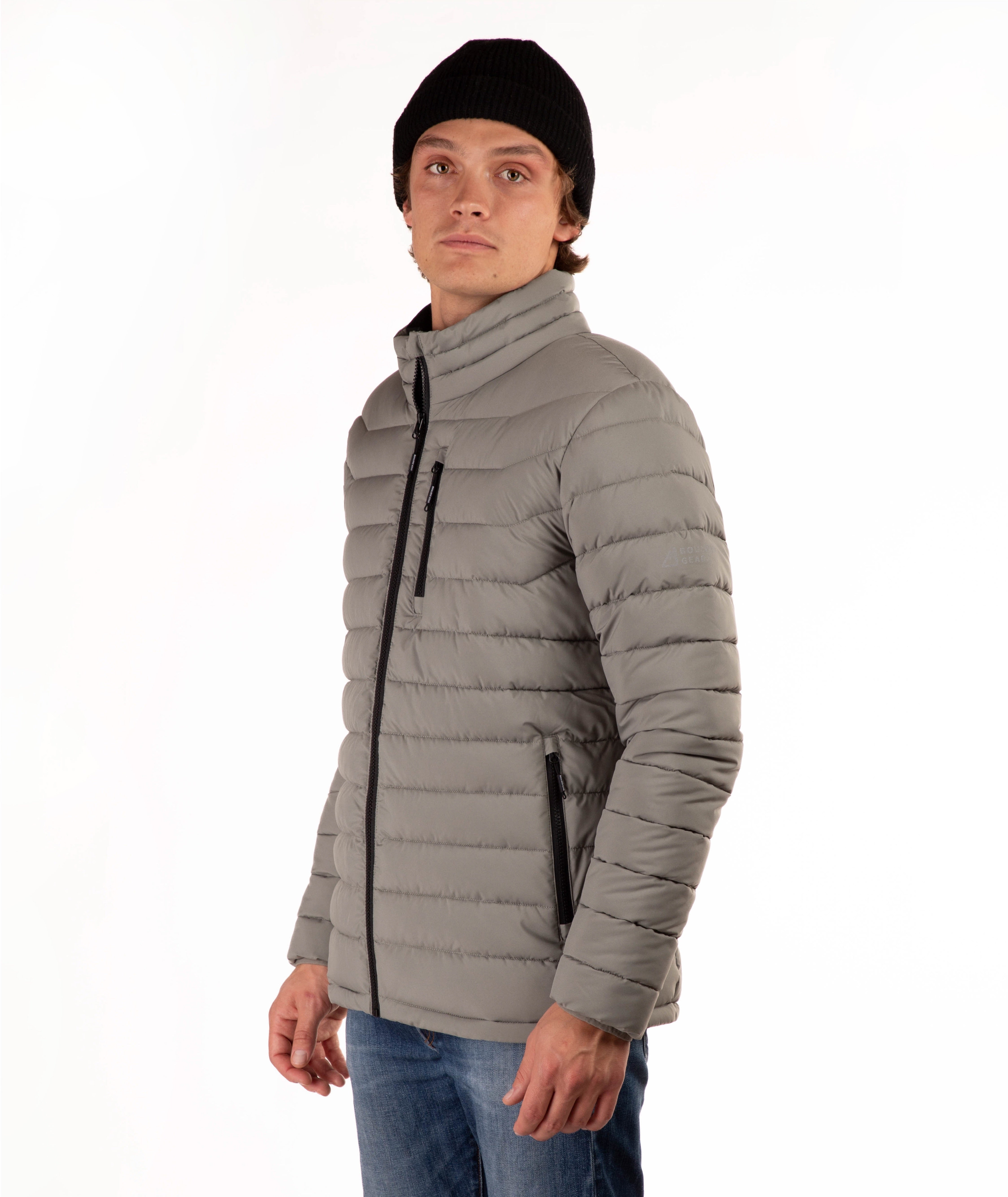 A man wearing a taupe quilted puffer jacket with a high collar, black zipper details, a black beanie, and blue jeans, standing against a plain white background.