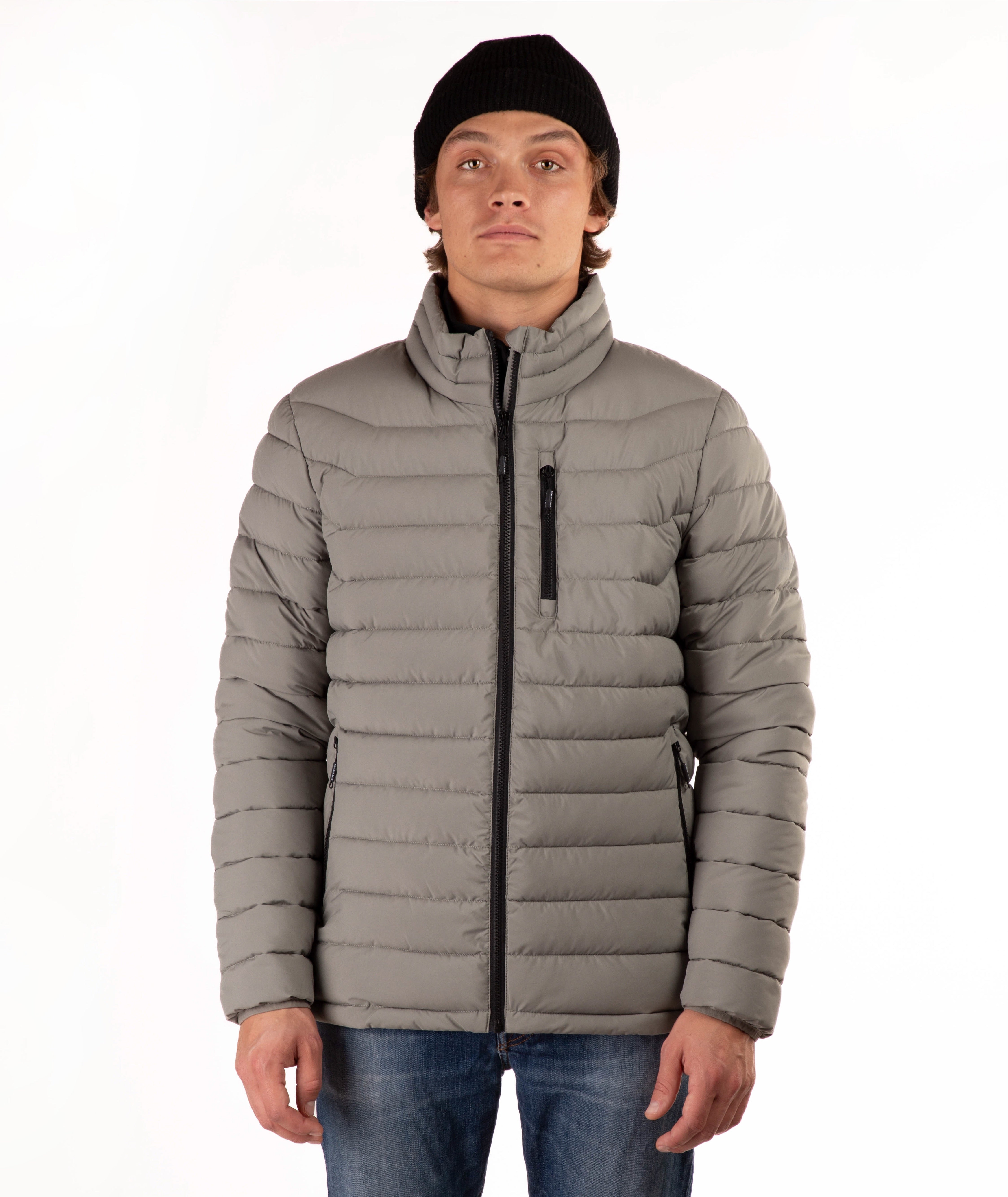 A man wearing a taupe quilted puffer jacket with a high collar, black zipper details, a black beanie, and blue jeans, standing against a plain white background.