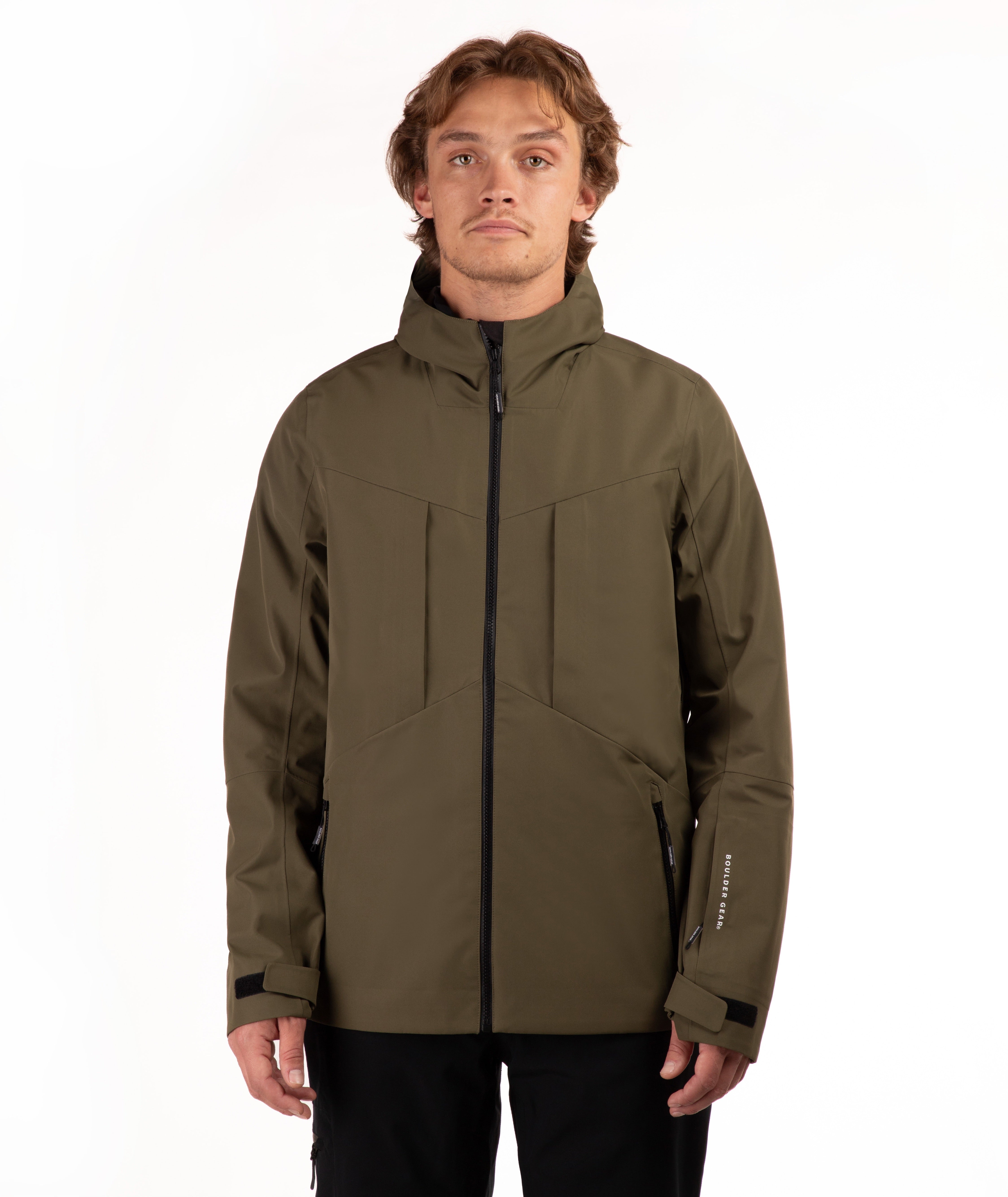 A man wearing an olive green technical outdoor jacket with black zippers and black pants, standing against a plain white studio background.